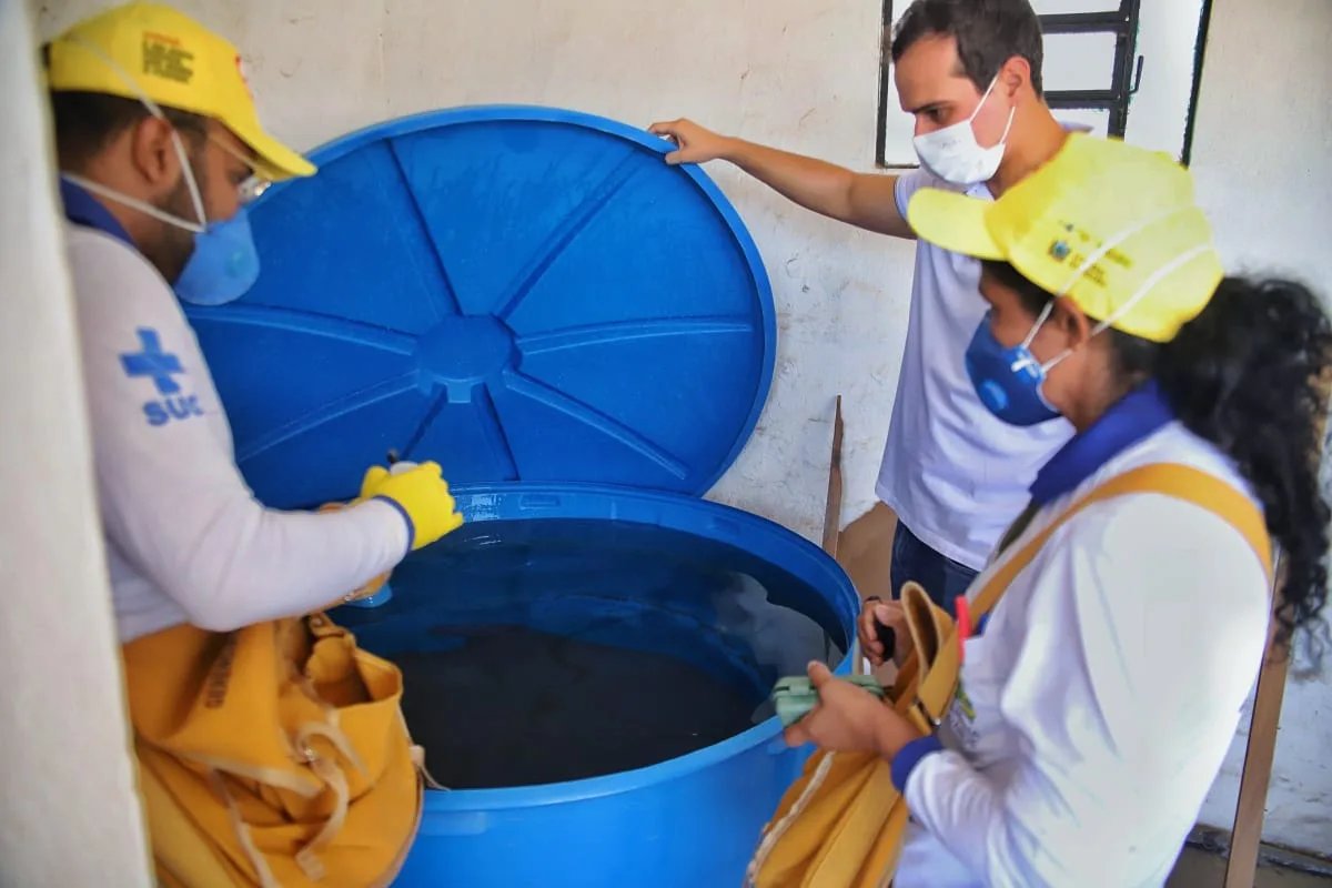 Agentes de saúde de boné amarelo e colete do SUS inspecionam uma caixa d'água azul aberta em uma residência, procurando focos do mosquito.