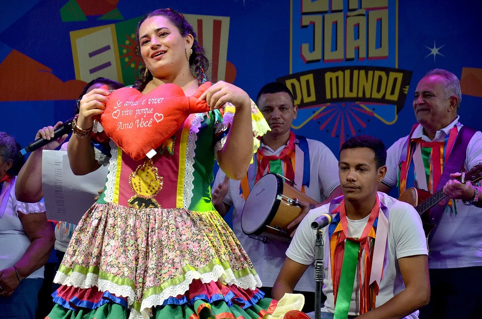 Mulher com vestido junino colorido segura um coração de tecido no palco, cercada por músicos com instrumentos, sob painel do São João.