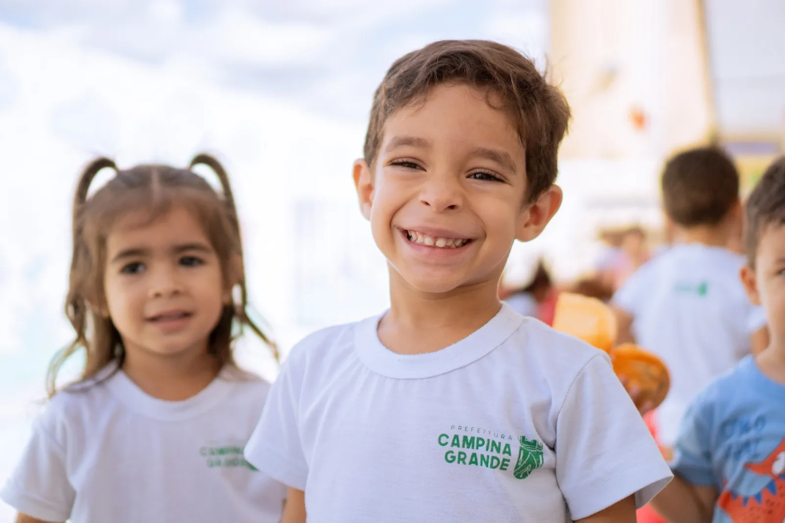 Duas crianças sorriem usando camisetas brancas com o brasão da Prefeitura de Campina Grande, em um ambiente escolar.
