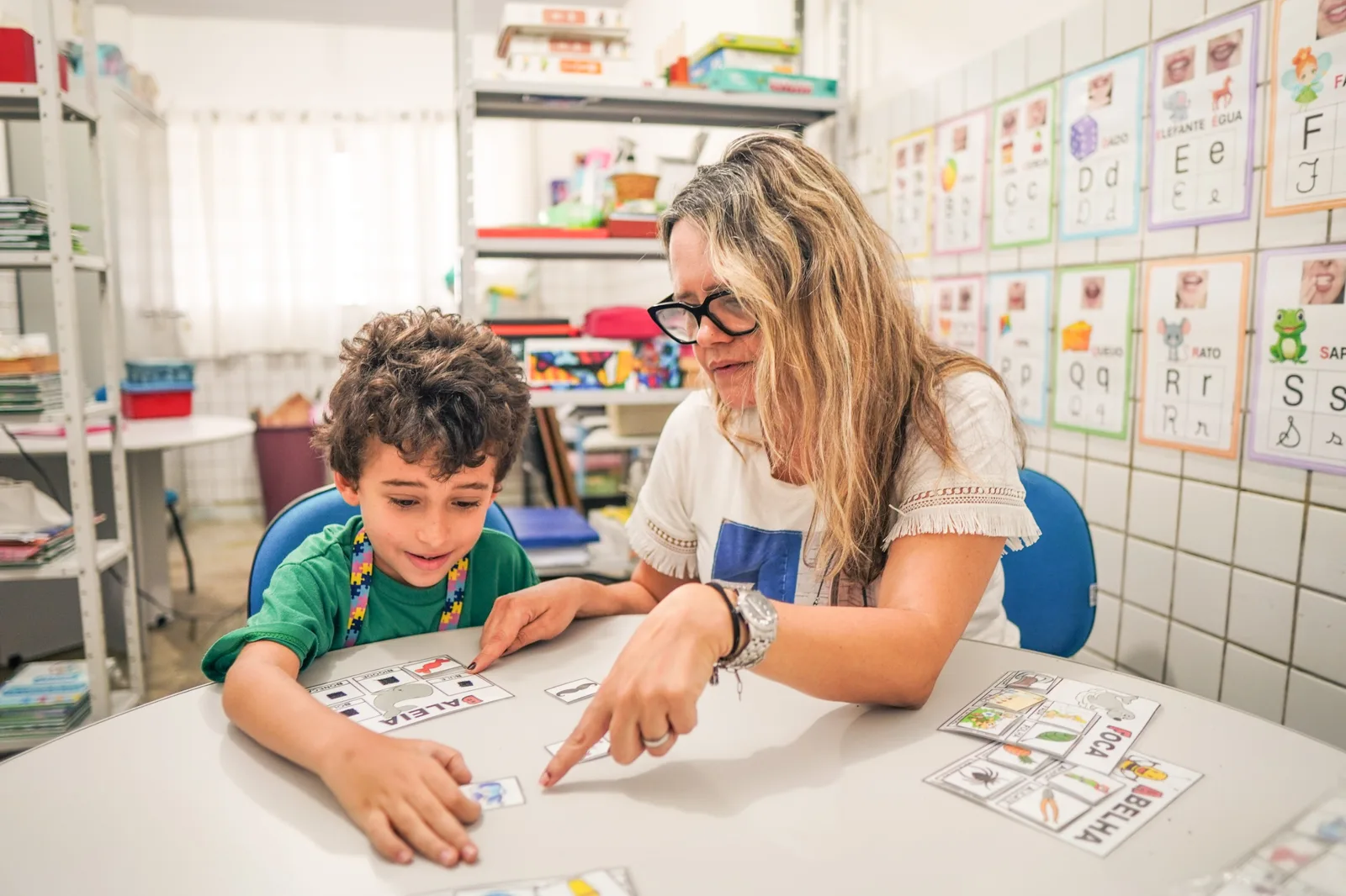 Professora aponta para cartões de letras sobre uma mesa enquanto um menino de camiseta verde acompanha a atividade, em sala de alfabetização com parede de alfabeto ilustrado.