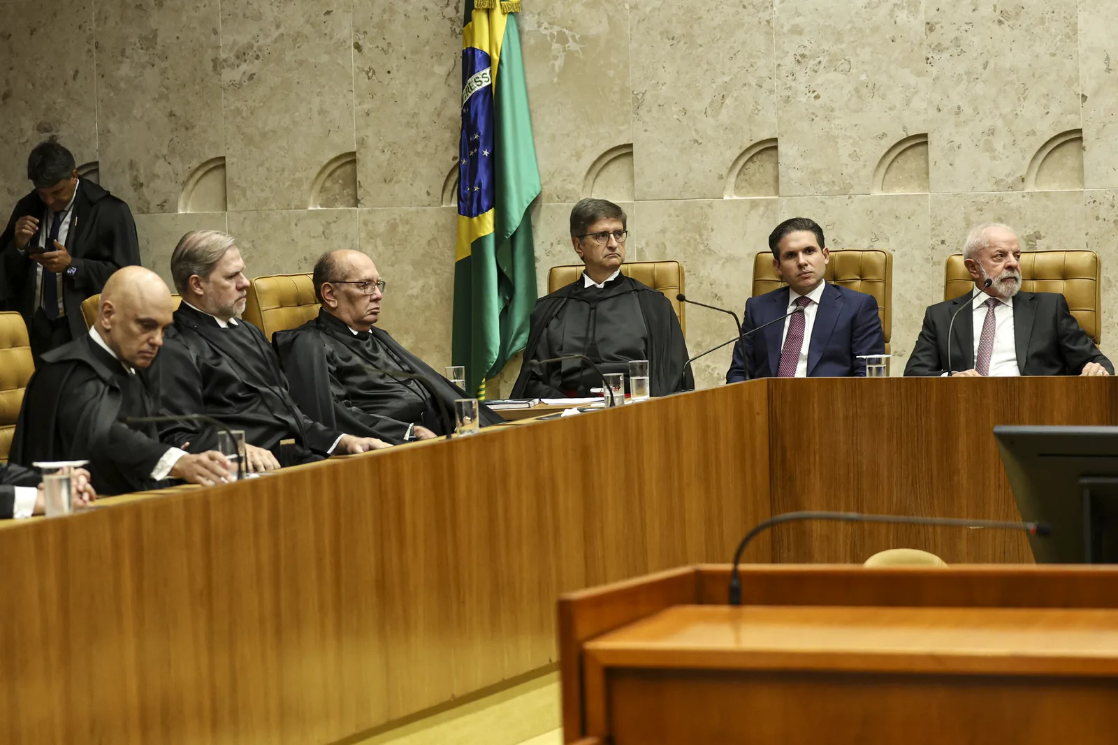 Ministros do Supremo Tribunal Federal sentados de toga na bancada do plenário, com a bandeira do Brasil ao fundo e microfones sobre a mesa de madeira.