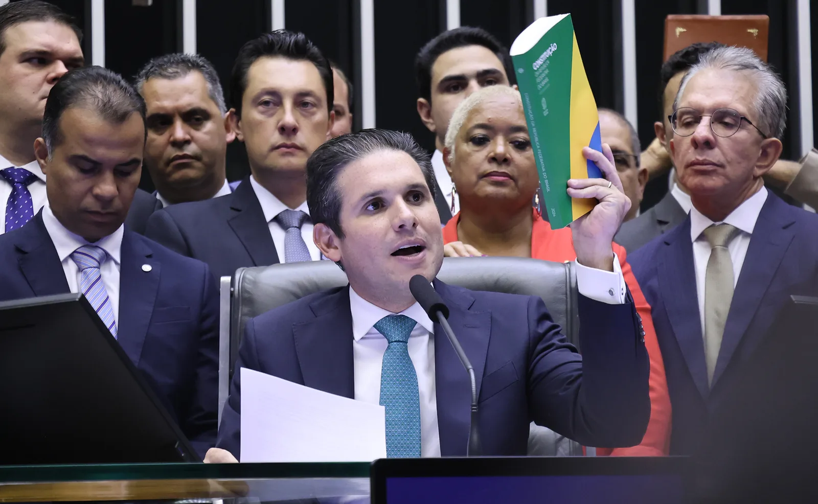 Hugo Motta, de terno e gravata azul, discursa na mesa diretora da Câmara segurando um exemplar da Constituição, cercado por parlamentares.
