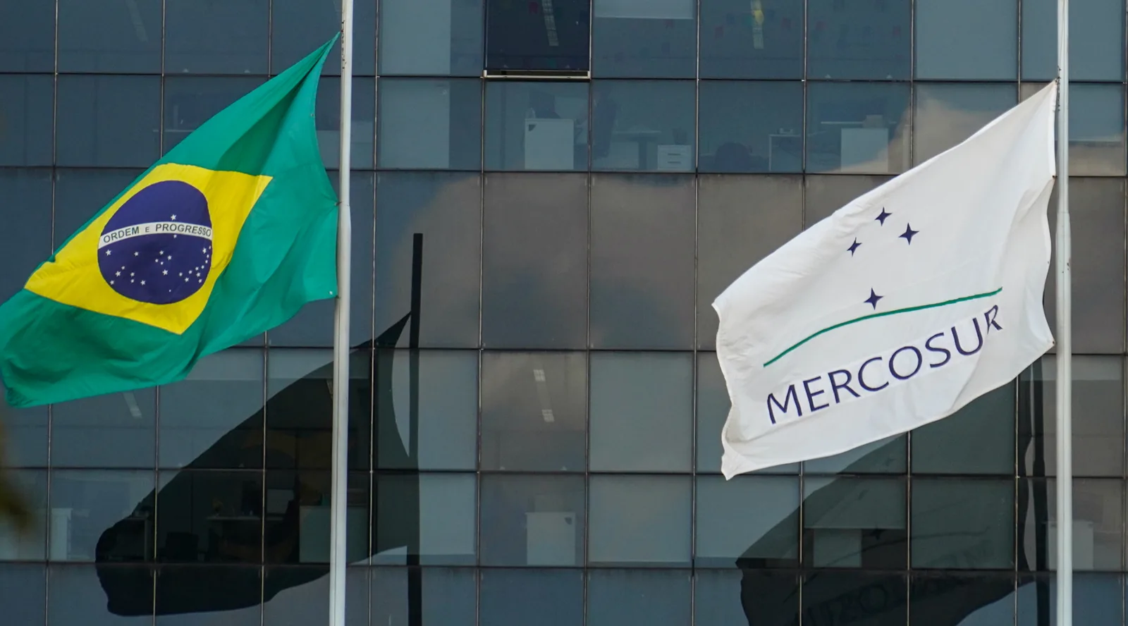 Bandeiras do Brasil e do Mercosul hasteadas diante de um edifício.