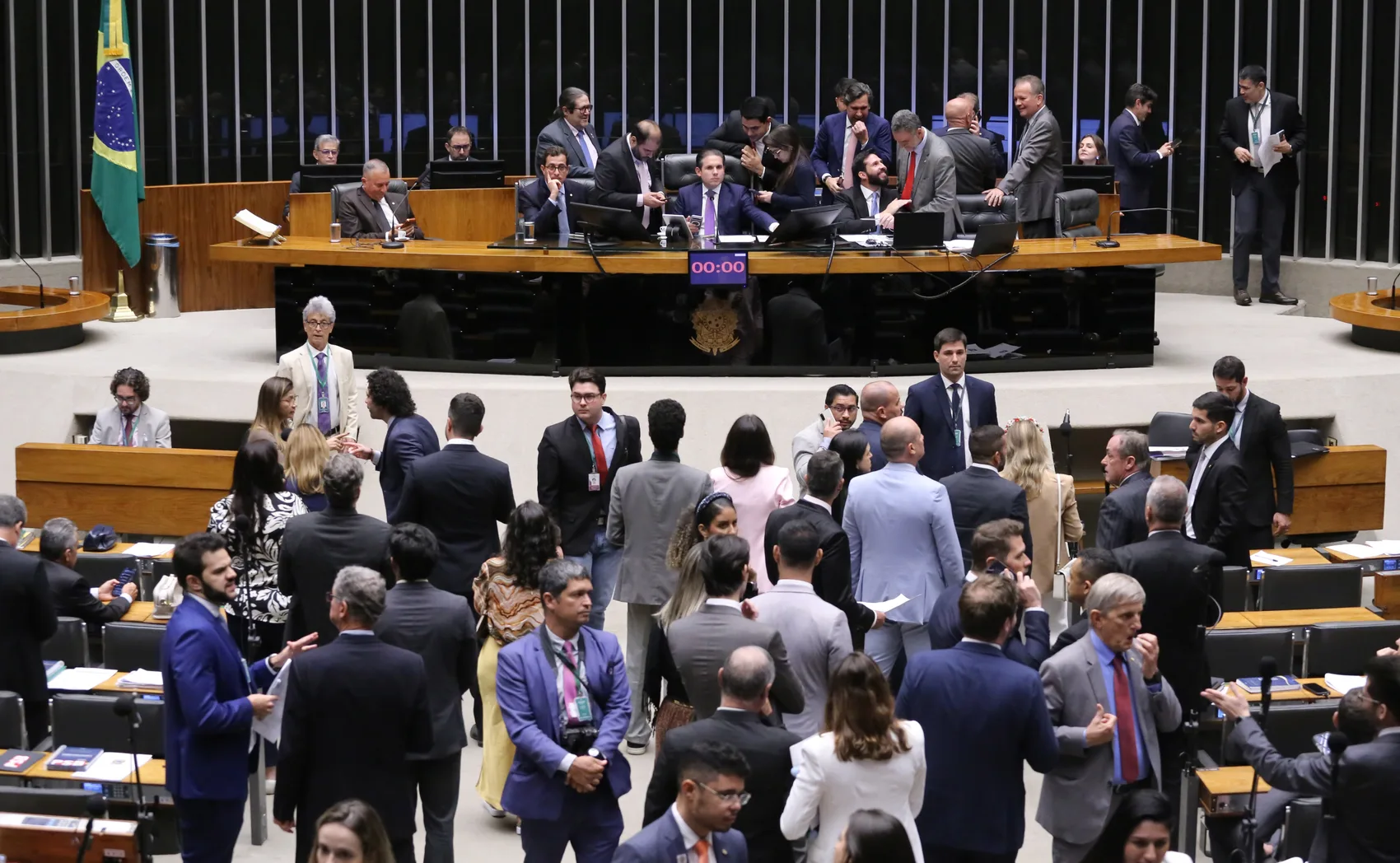 Vista ampla do plenário da Câmara dos Deputados, com a mesa diretora ao fundo, a bandeira do Brasil e dezenas de parlamentares reunidos entre as bancadas.