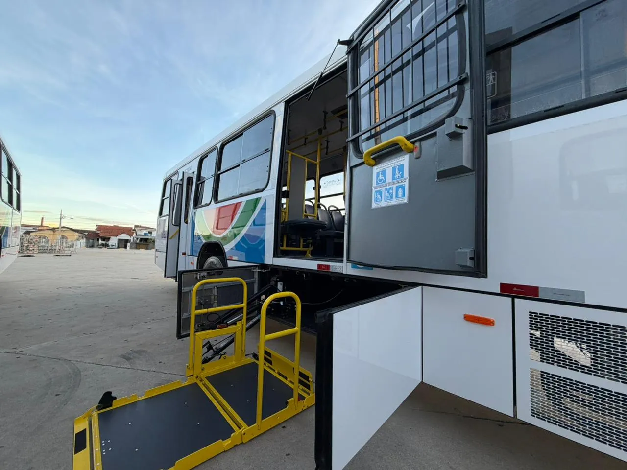 Ônibus novos brancos enfileirados em um pátio ao entardecer, com a plataforma elevatória para cadeirantes aberta em um dos veículos, em Campina Grande.