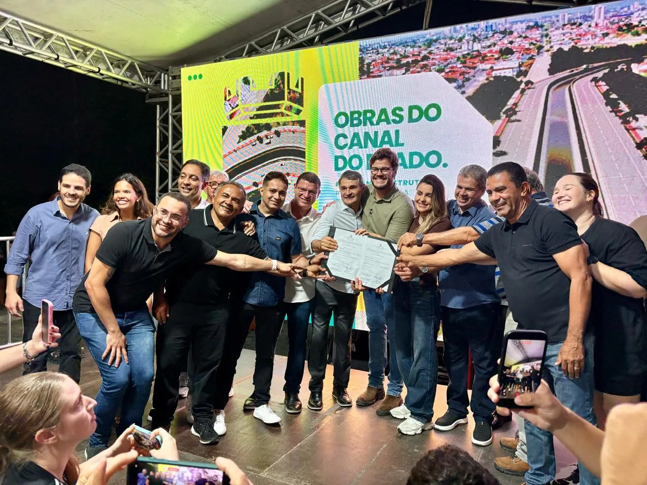 Autoridades e moradores seguram a ordem de serviço das obras do Canal do Prado em um palco, com painel do projeto ao fundo, em Campina Grande.