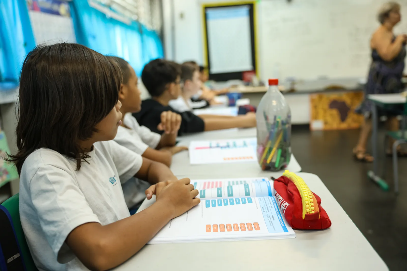 Alunos sentados em fileira de carteiras acompanham a aula com o material didático aberto, em sala de aula.
