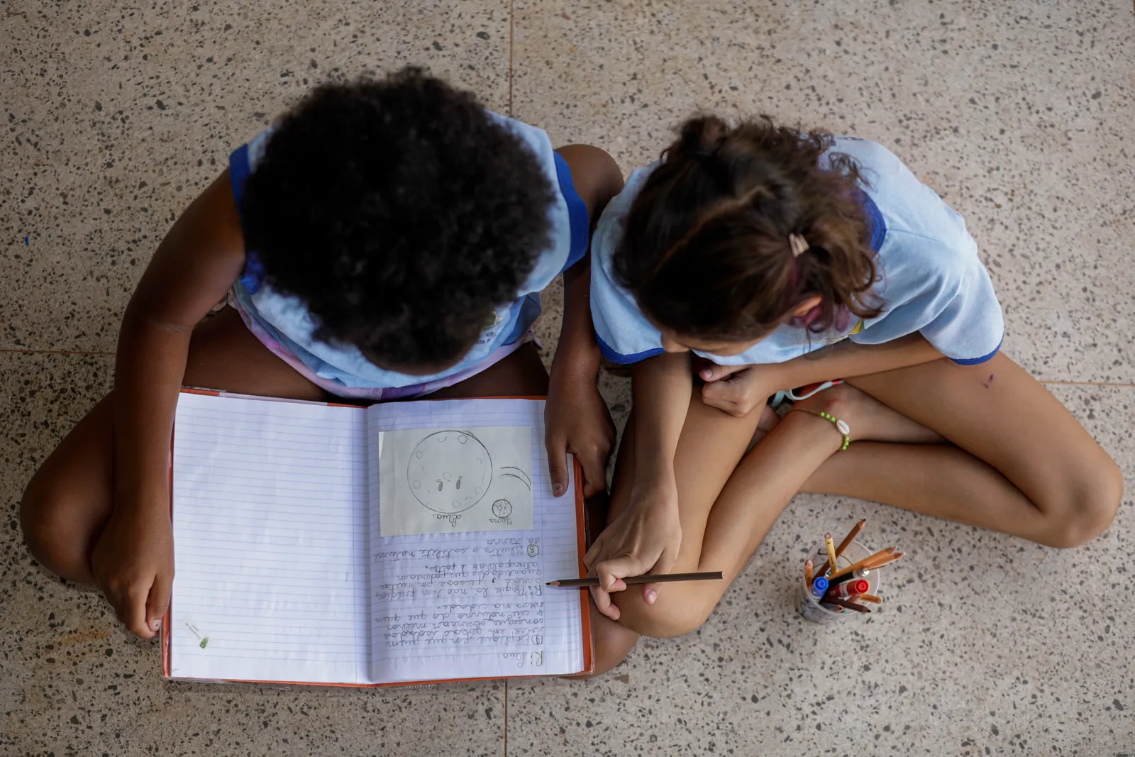 Vista de cima de duas estudantes de escola pública sentadas no chão, com caderno aberto, lápis e lápis de cor ao lado