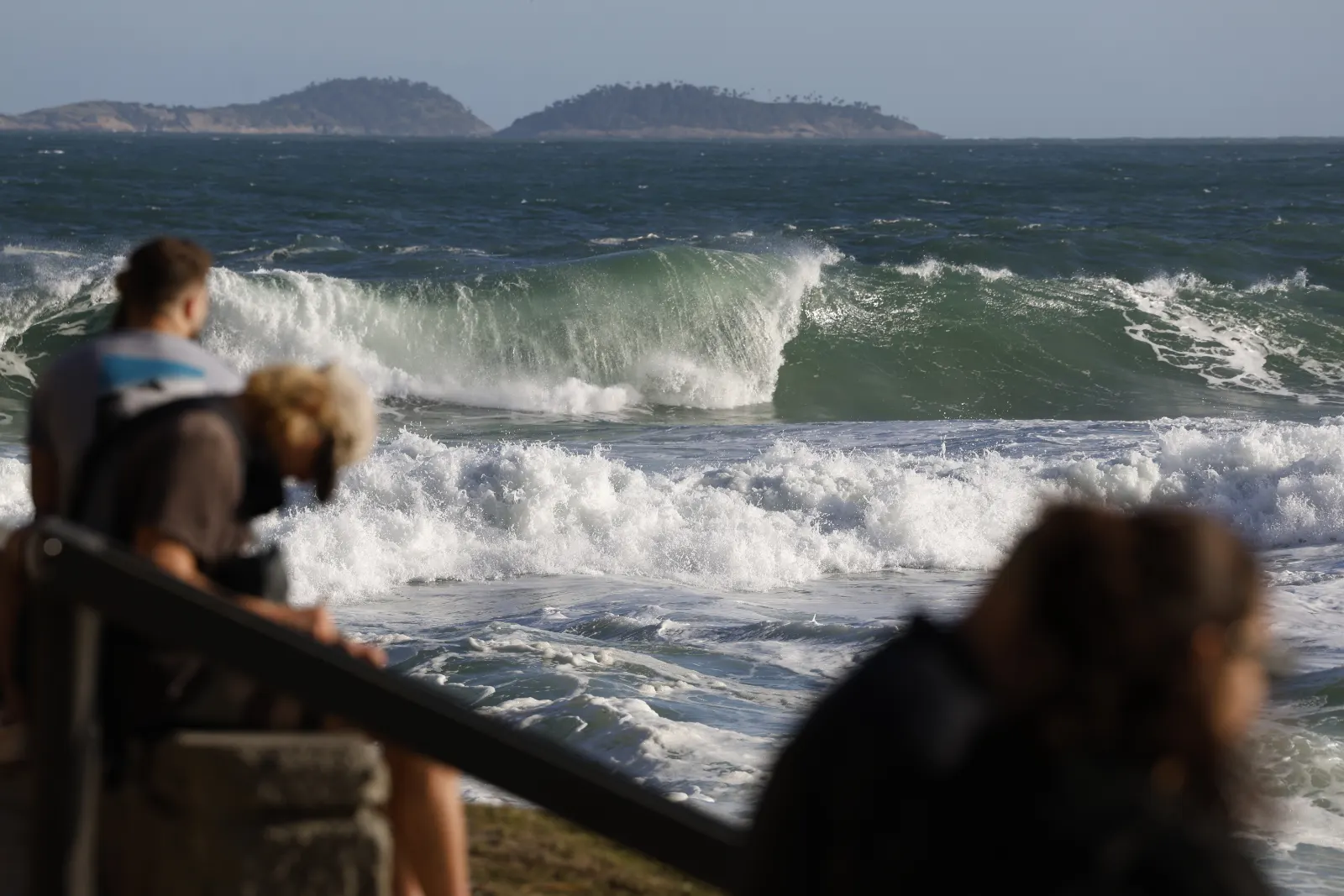 Ondas grandes de ressaca quebram junto à orla enquanto pessoas observam o mar agitado sob céu claro.