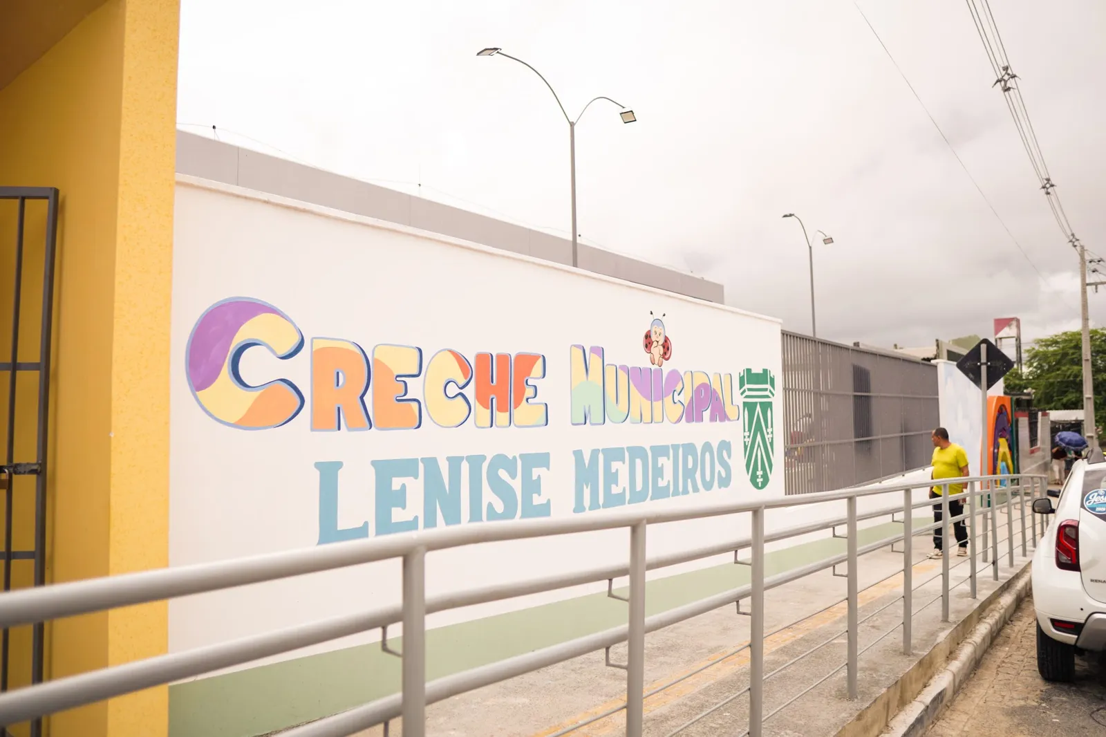 Fachada branca e amarela da Creche Municipal Lenise Medeiros, com o nome pintado na parede e rampa de acesso com corrimão, no bairro das Malvinas, em Campina Grande.