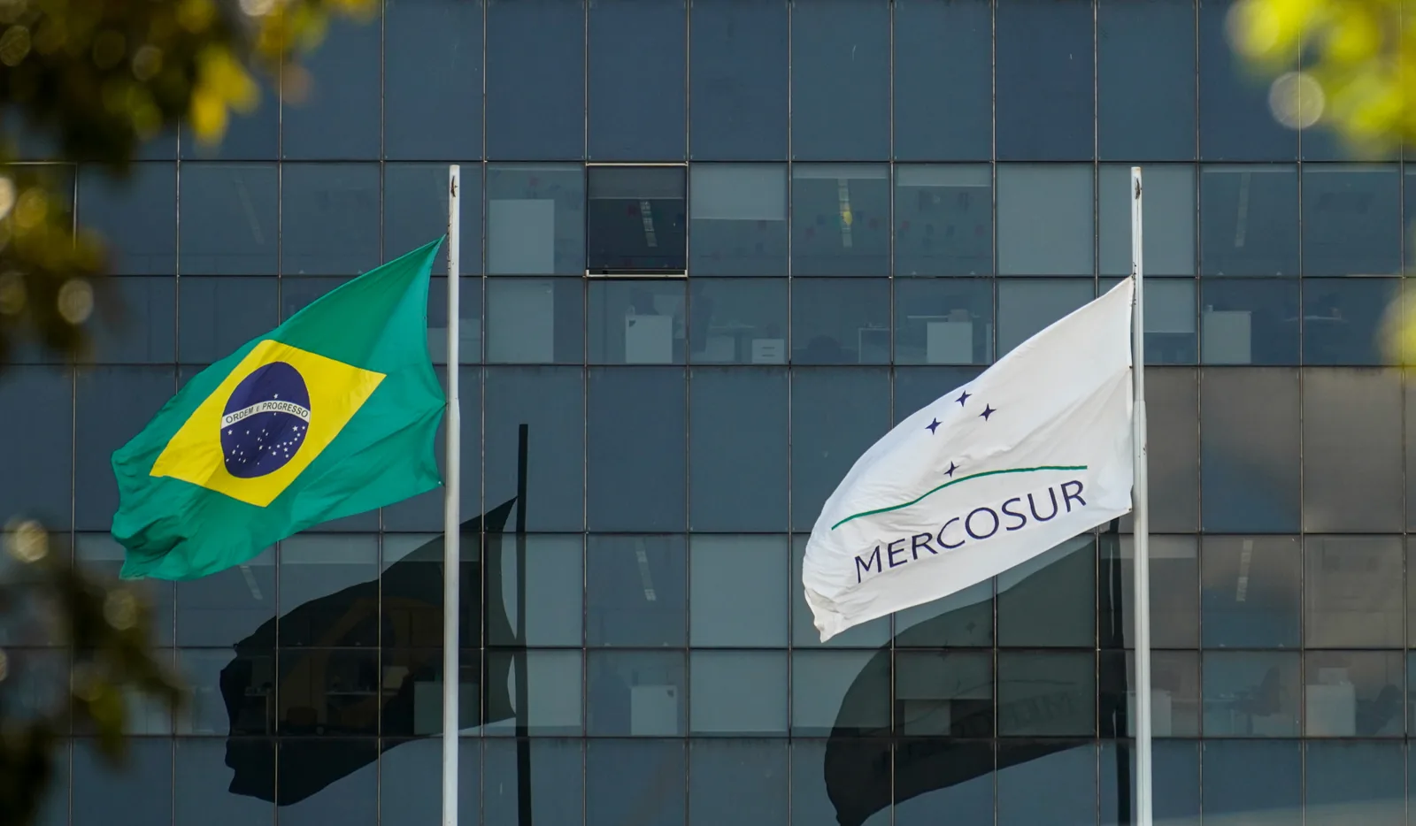 Bandeiras do Brasil e do Mercosul hasteadas diante de um prédio envidraçado em Brasília.