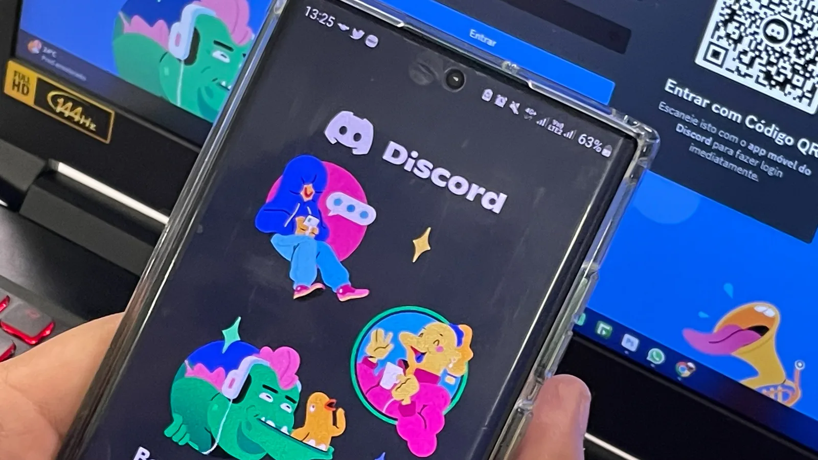 Mão segura celular com o aplicativo Discord aberto na tela de entrada, diante de um computador que exibe a página de login por código QR.
