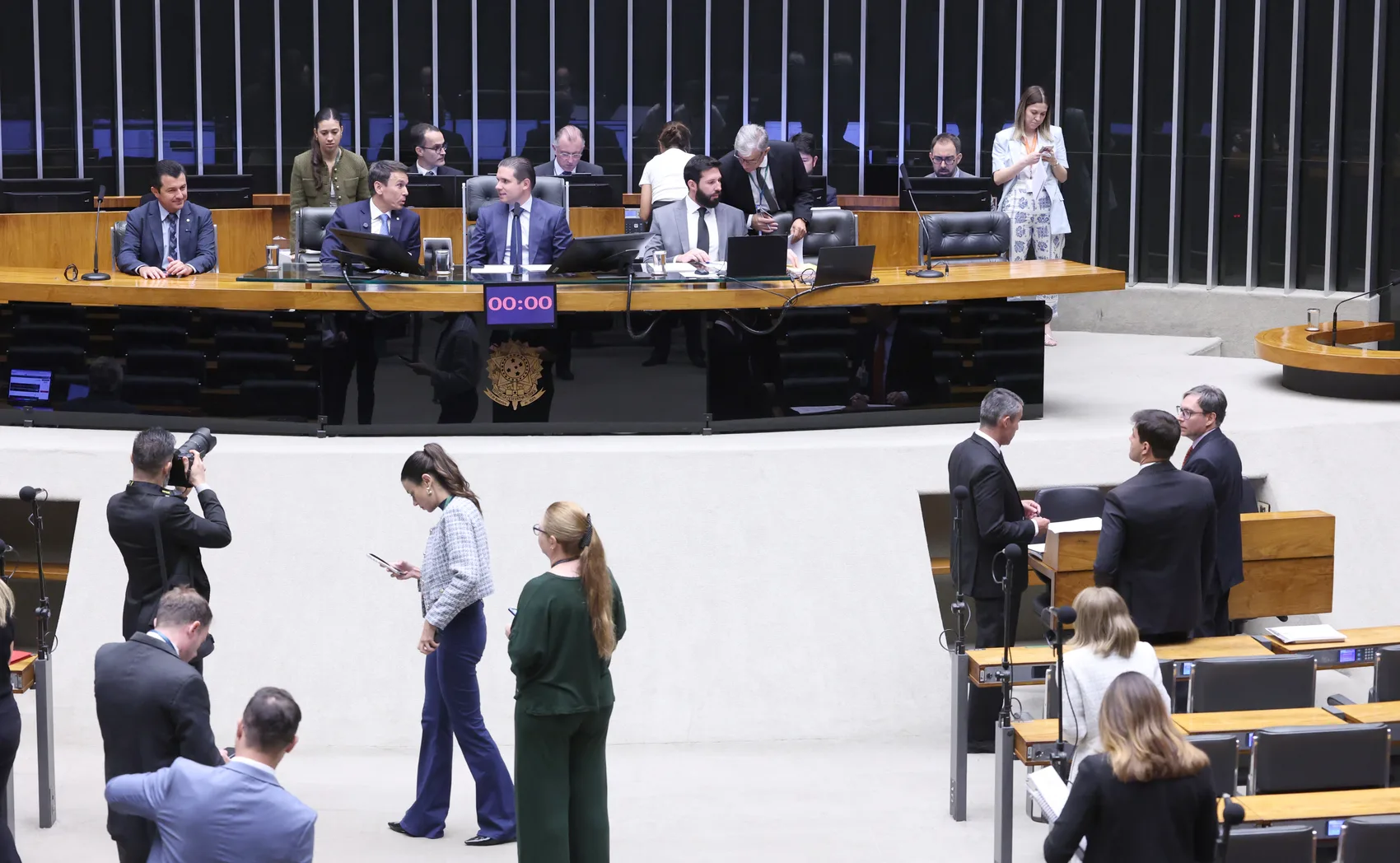 Vista ampla do plenário da Câmara dos Deputados, com a mesa diretora, o brasão da República e parlamentares reunidos antes de uma sessão.