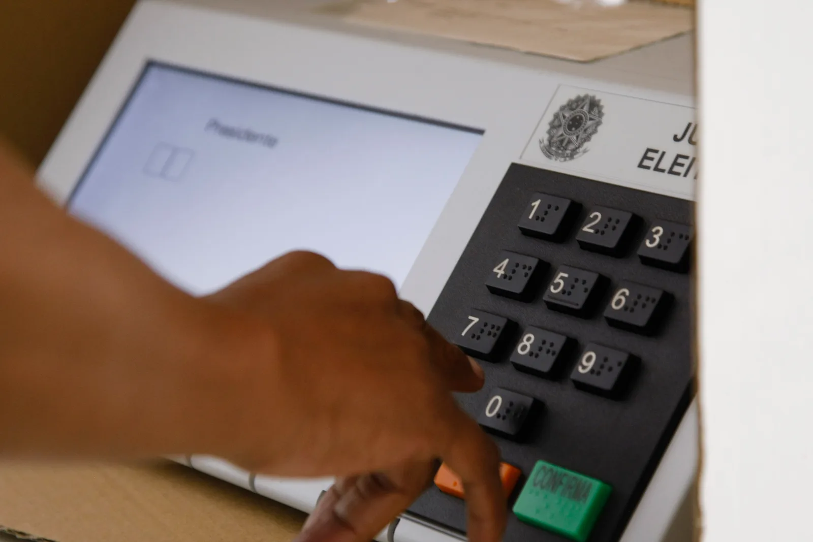 Mão de um eleitor pressiona as teclas numéricas da urna eletrônica, ao lado da tela que exibe a votação para presidente e do emblema da Justiça Eleitoral.