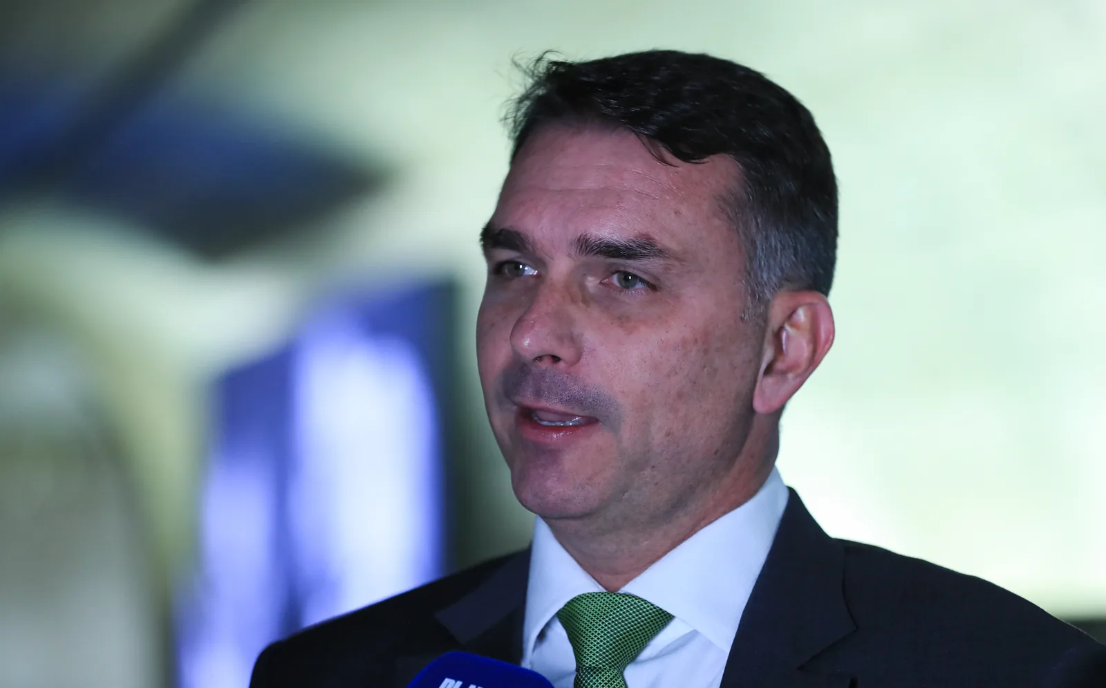 Flávio Bolsonaro, de terno escuro e gravata verde, fala a jornalistas diante de microfones.
