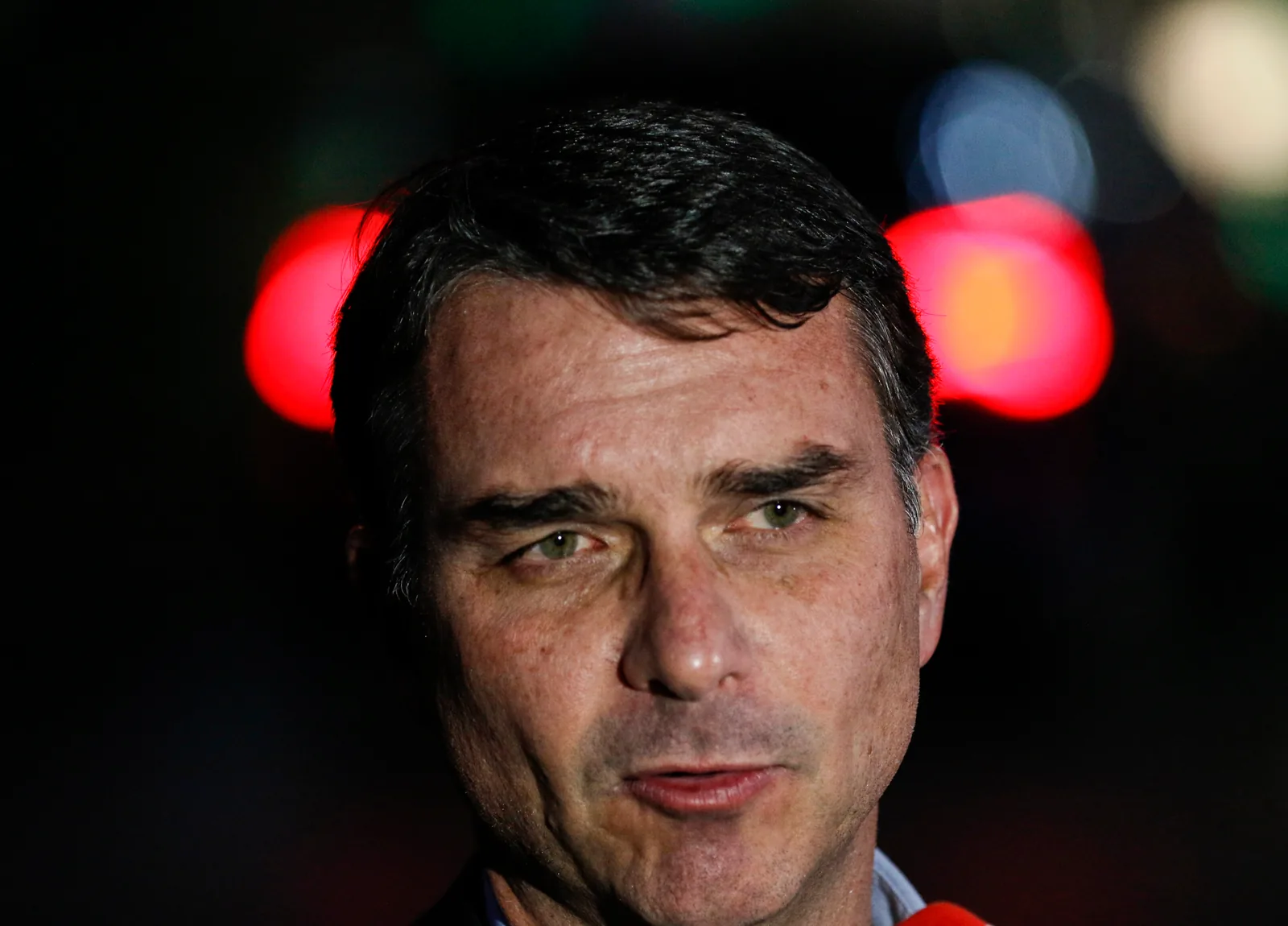 Rosto de Flávio Bolsonaro em close, falando a jornalistas à noite, com luzes desfocadas ao fundo.