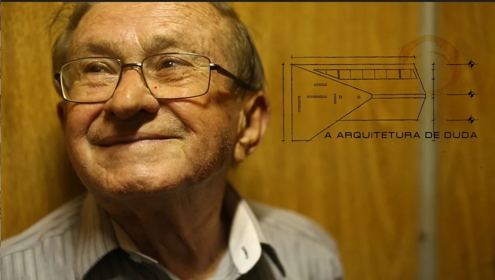 Geraldino Duda sorrindo, de óculos, ao lado de um painel com o desenho do Teatro Municipal e os dizeres A Arquitetura de Duda.