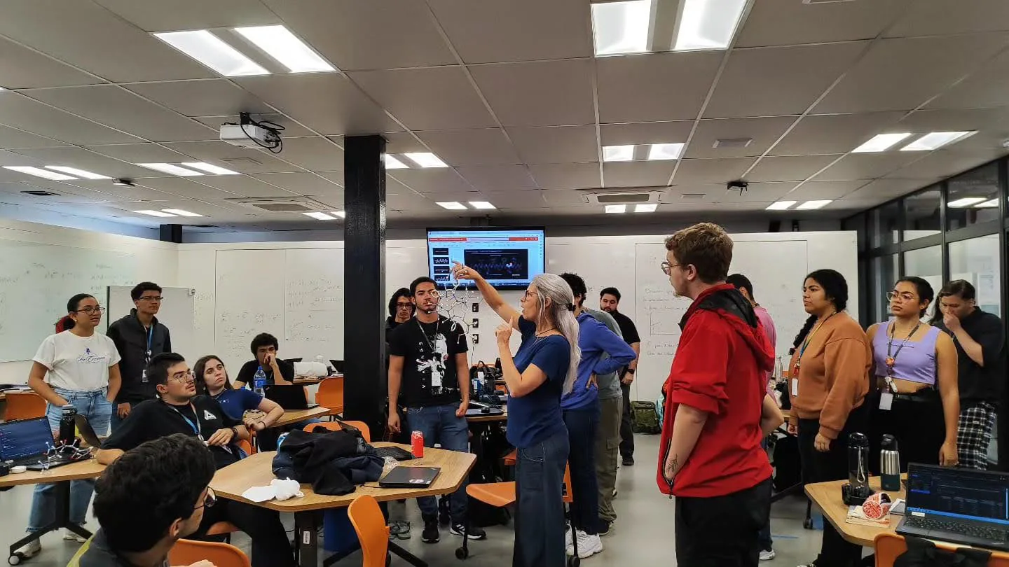 Estudantes universitários reunidos em sala de aula com notebooks abertos sobre as mesas, diante de uma tela com apresentação.