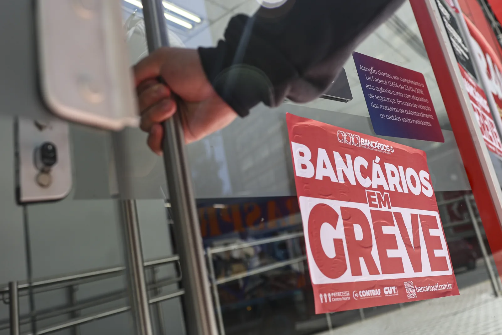 Cartaz vermelho com os dizeres Bancários em Greve colado no vidro da porta de uma agência, enquanto uma mão puxa a maçaneta.