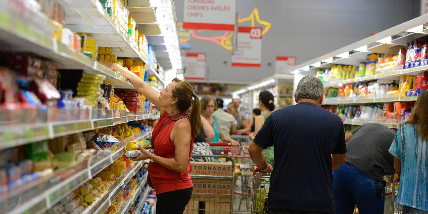 Consumidor observa frutas e hortaliças expostas em banca de supermercado, com etiquetas de preço à mostra.