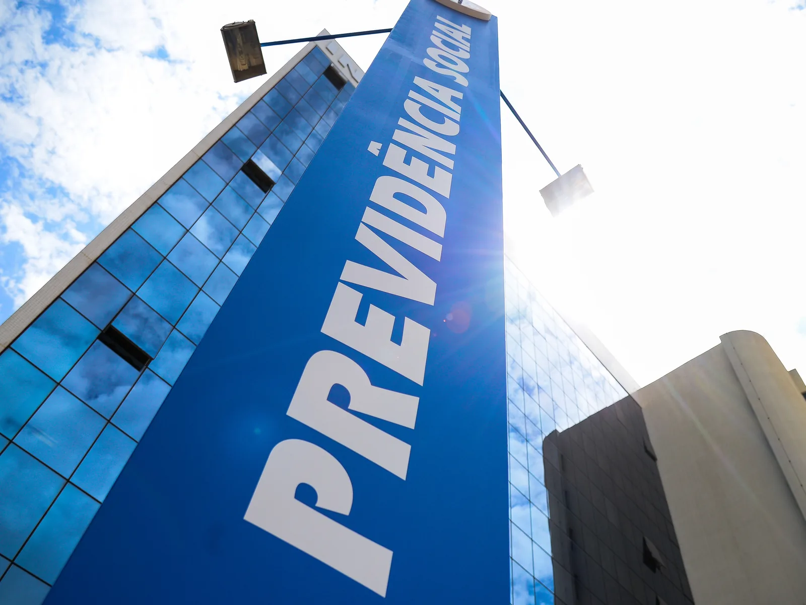 Fachada de prédio espelhado com uma faixa vertical azul de ponta a ponta trazendo a inscrição Previdência Social, fotografada de baixo para cima contra o céu.