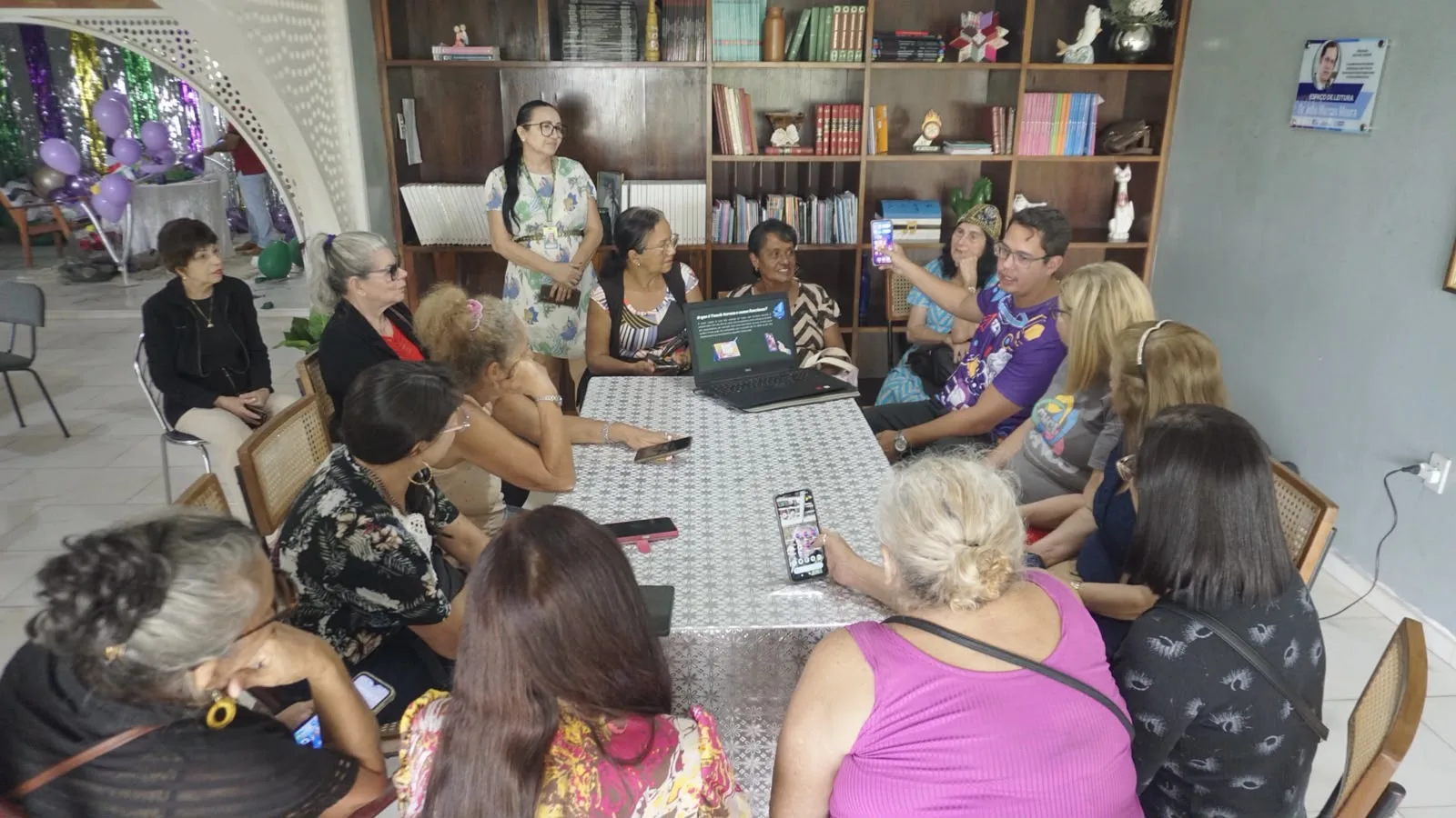 Roda de pessoas idosas sentadas em volta de uma mesa com notebook e celulares, durante minicurso de letramento digital em espaço comunitário.