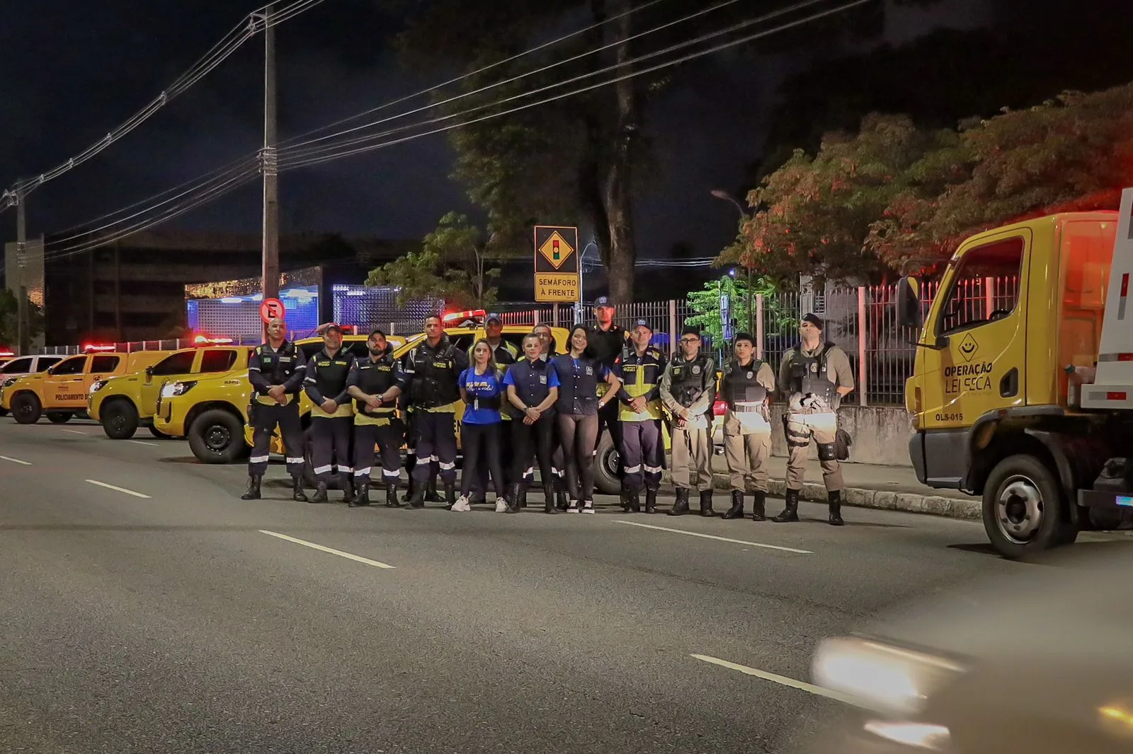 Agentes do Detran-PB e policiais militares perfilados numa avenida à noite, diante de viaturas amarelas e de um caminhão com a inscrição Operação Lei Seca.