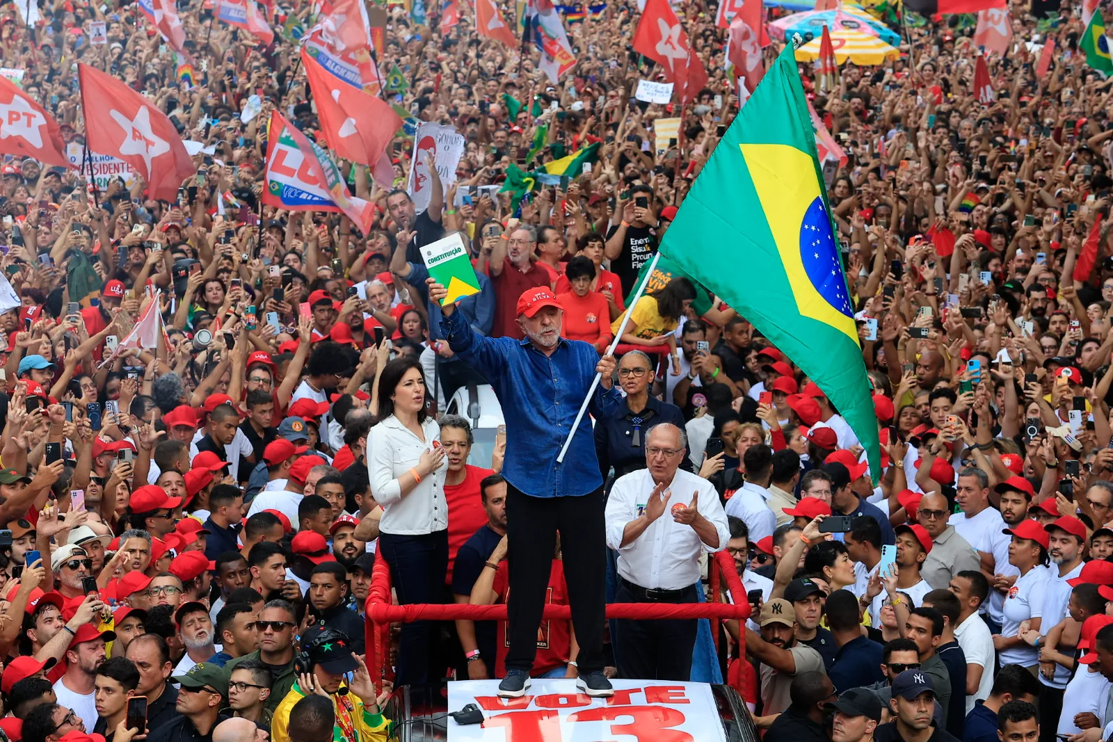 Lula, de camisa azul, ergue uma bandeira do Brasil sobre um carro de som cercado por milhares de apoiadores com bandeiras vermelhas.