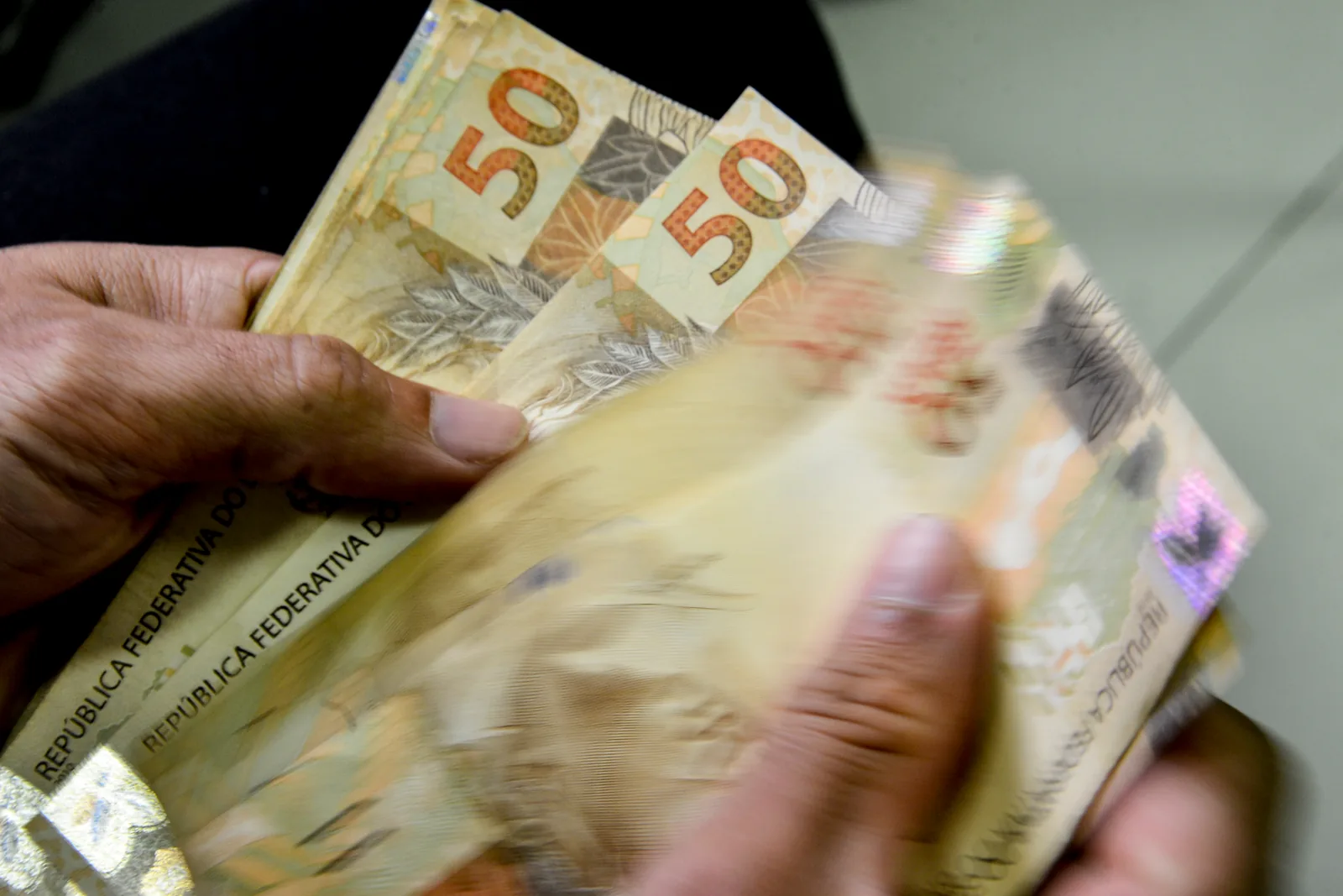 Mãos contam cédulas de cinquenta reais, com notas de real desfocadas ao fundo.