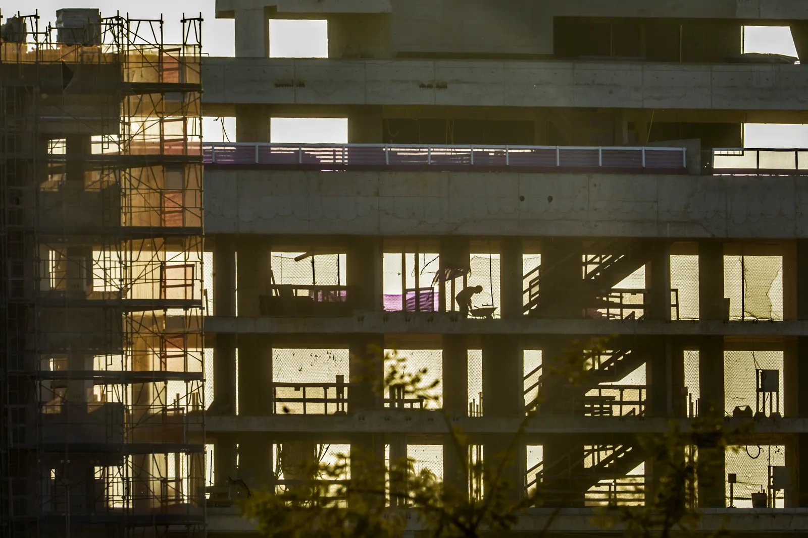 Prédios residenciais em construção ao entardecer, com andaimes, lajes vazadas e um operário em silhueta contra a luz.