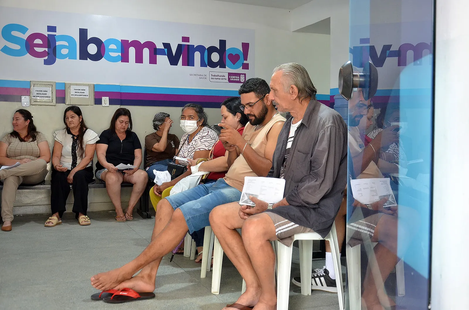 Pessoas sentadas na sala de espera de uma unidade de saúde de Campina Grande, com painel escrito Seja bem-vindo ao fundo.