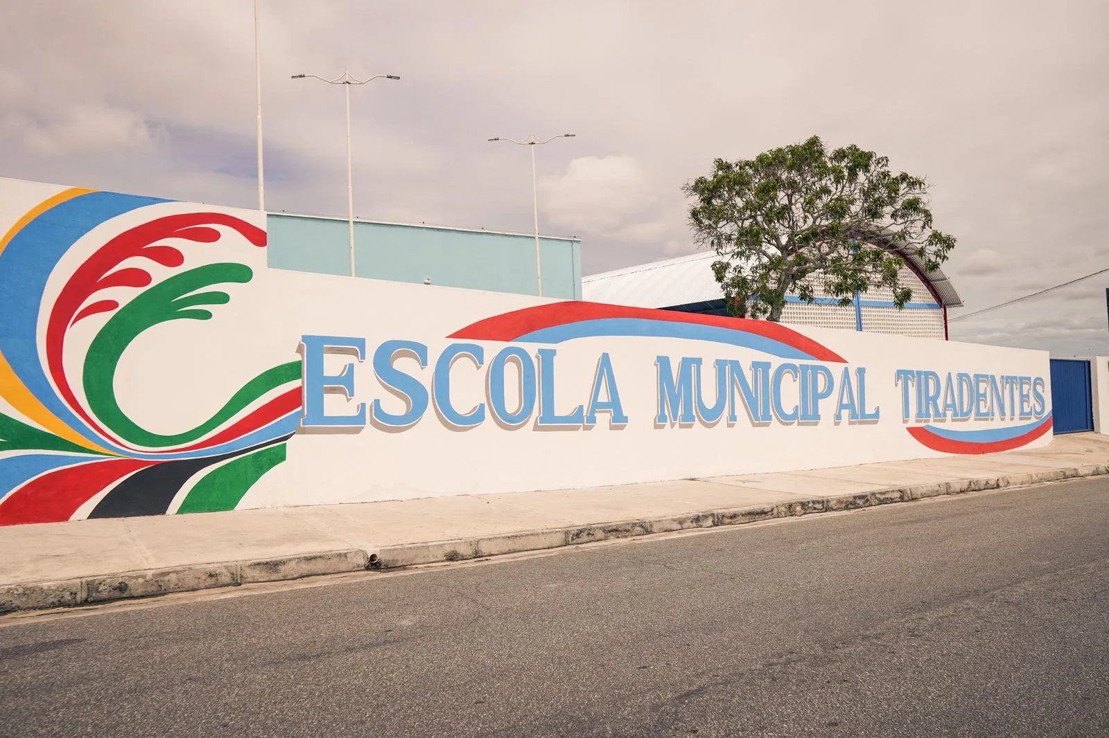 Muro branco da Escola Municipal Tiradentes com o nome em letras azuis e grafismo colorido na lateral, à beira de uma rua asfaltada.