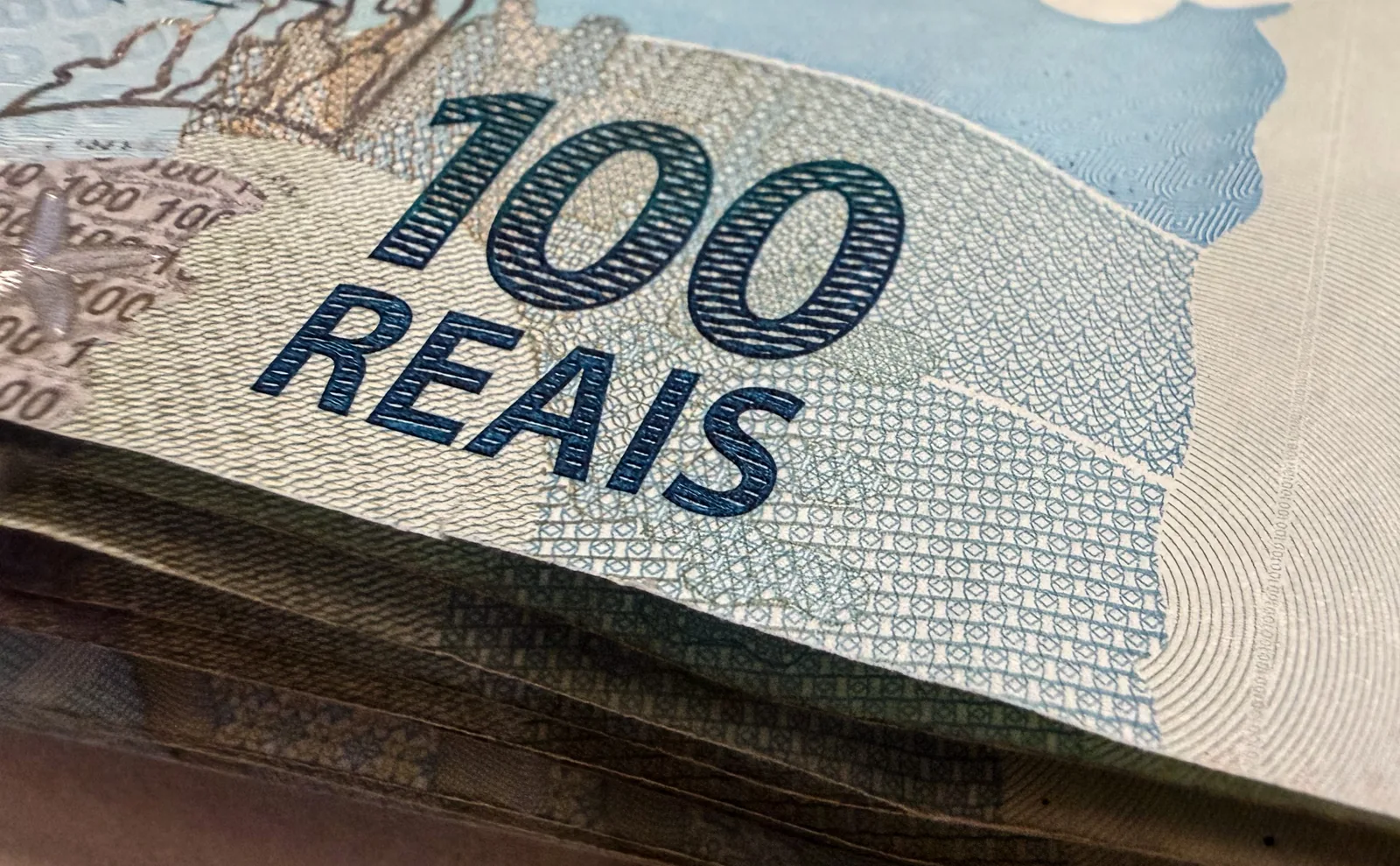Cédulas de cem reais empilhadas em close, com os dizeres 100 Reais em destaque.
