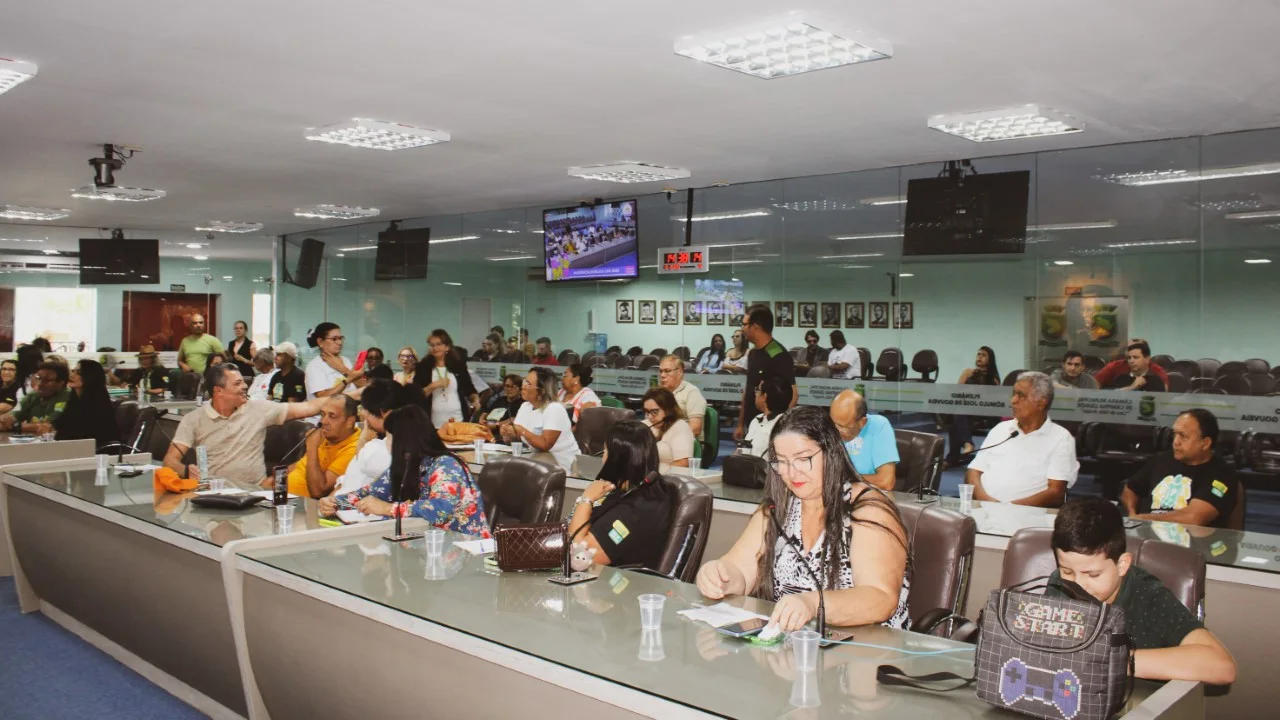 Público sentado nas bancadas do plenário da Câmara Municipal de Campina Grande durante audiência pública, com telão exibindo a sessão ao fundo.