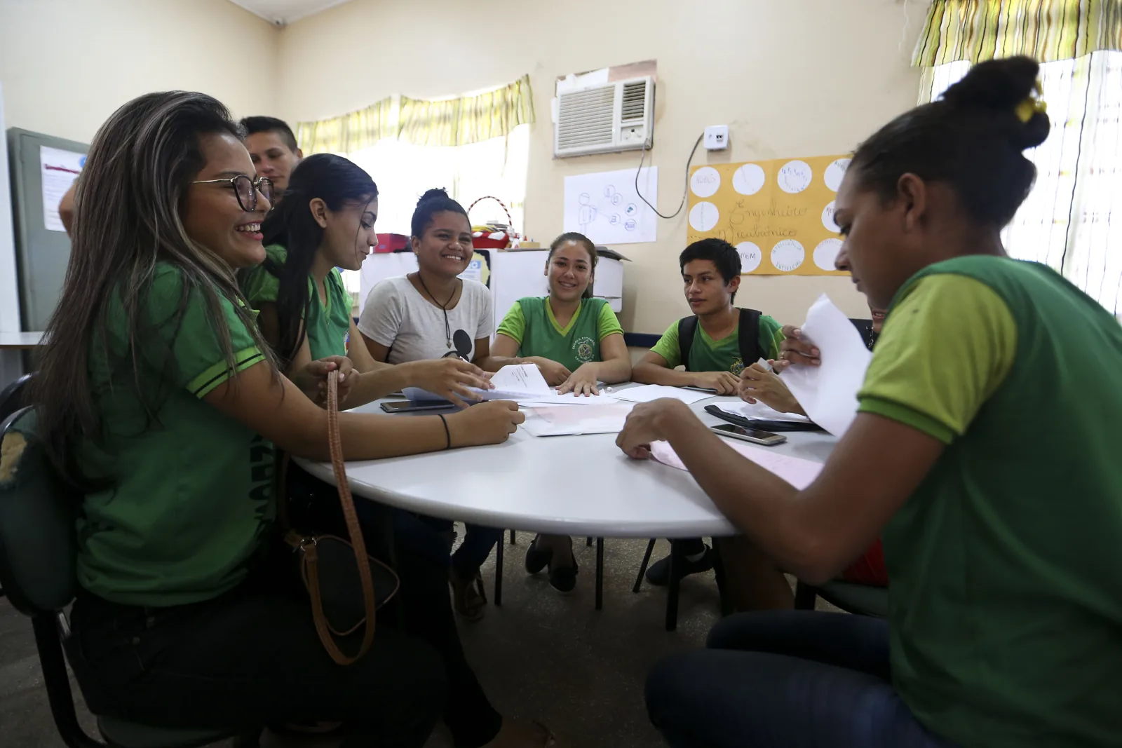 Seis estudantes de uniforme verde sentados em volta de uma mesa redonda numa sala de aula, conversando sobre folhas de papel.
