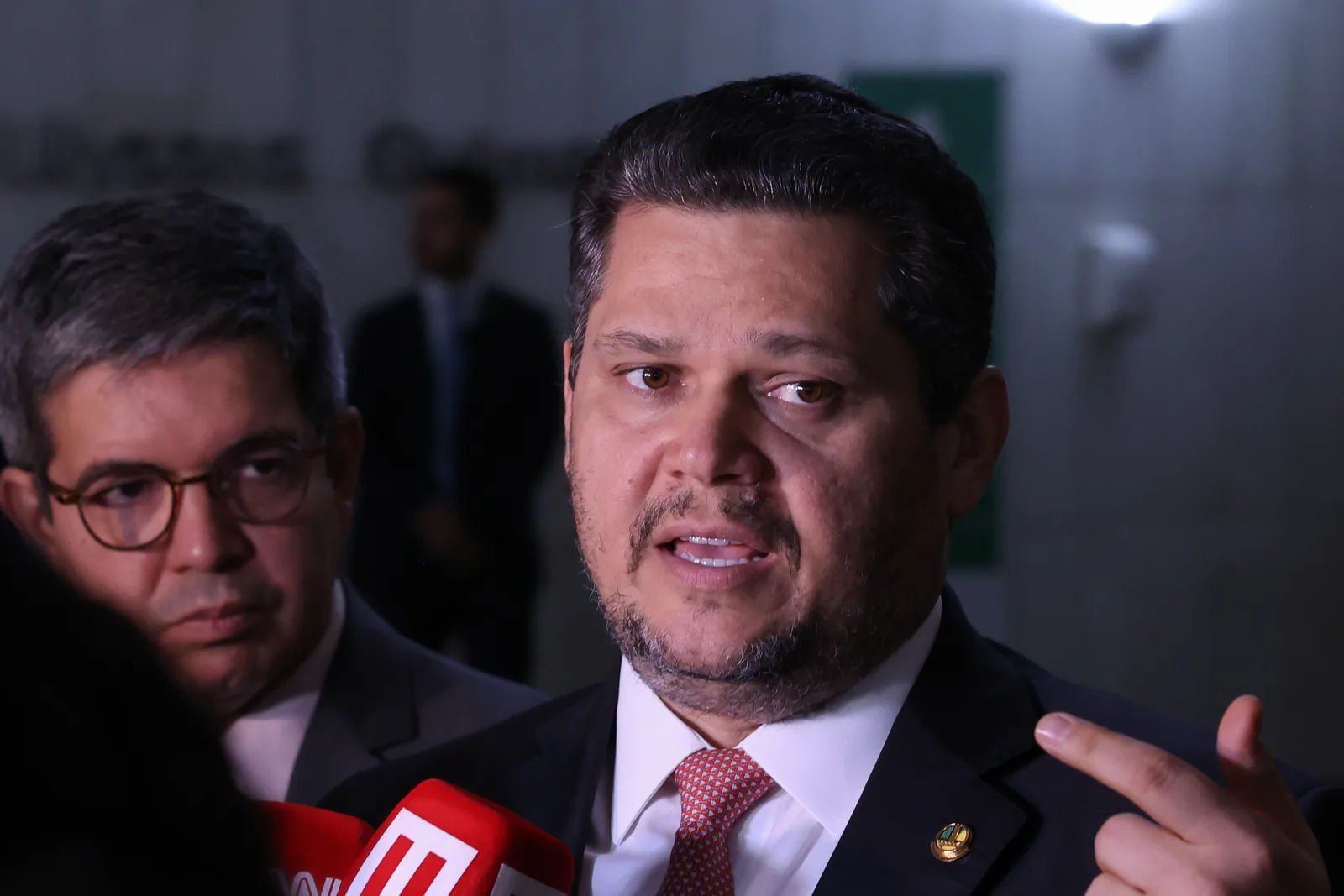 Davi Alcolumbre, de terno e gravata vermelha, fala a jornalistas diante de microfones.