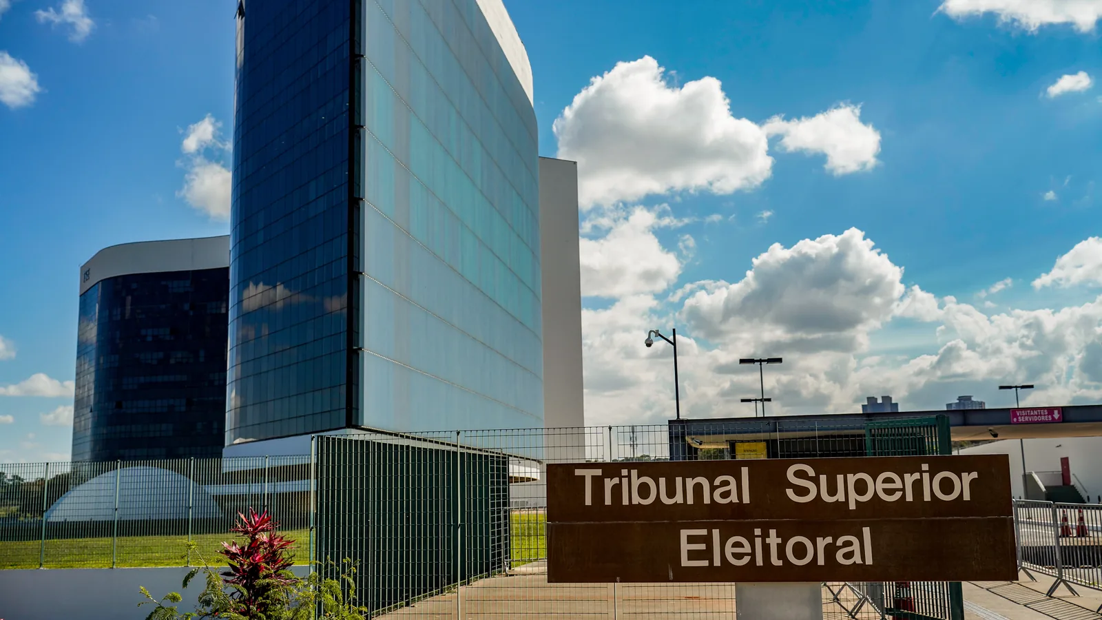 Placa de concreto com a inscrição Tribunal Superior Eleitoral em frente ao edifício-sede envidraçado da corte, em dia de céu azul com nuvens.