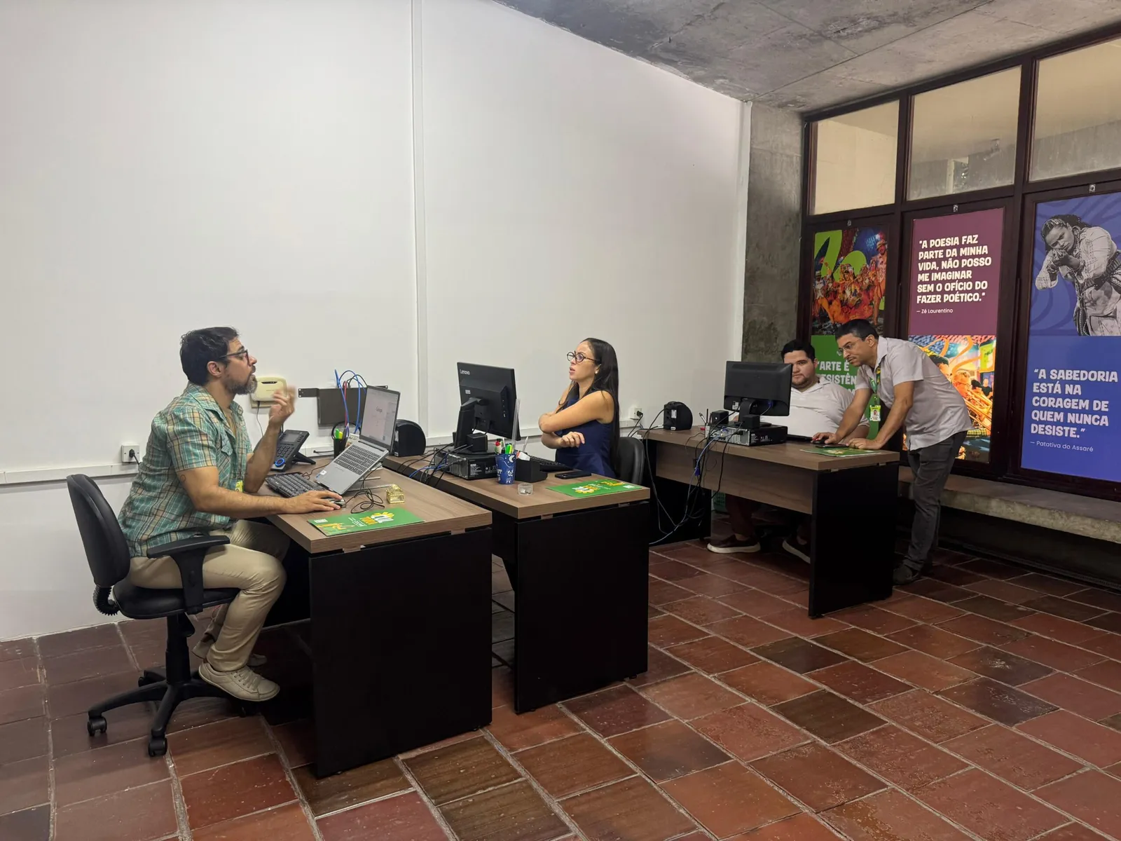 Espaço com três mesas, computadores e cartazes culturais na Sala do Fazedor de Cultura em Campina Grande.