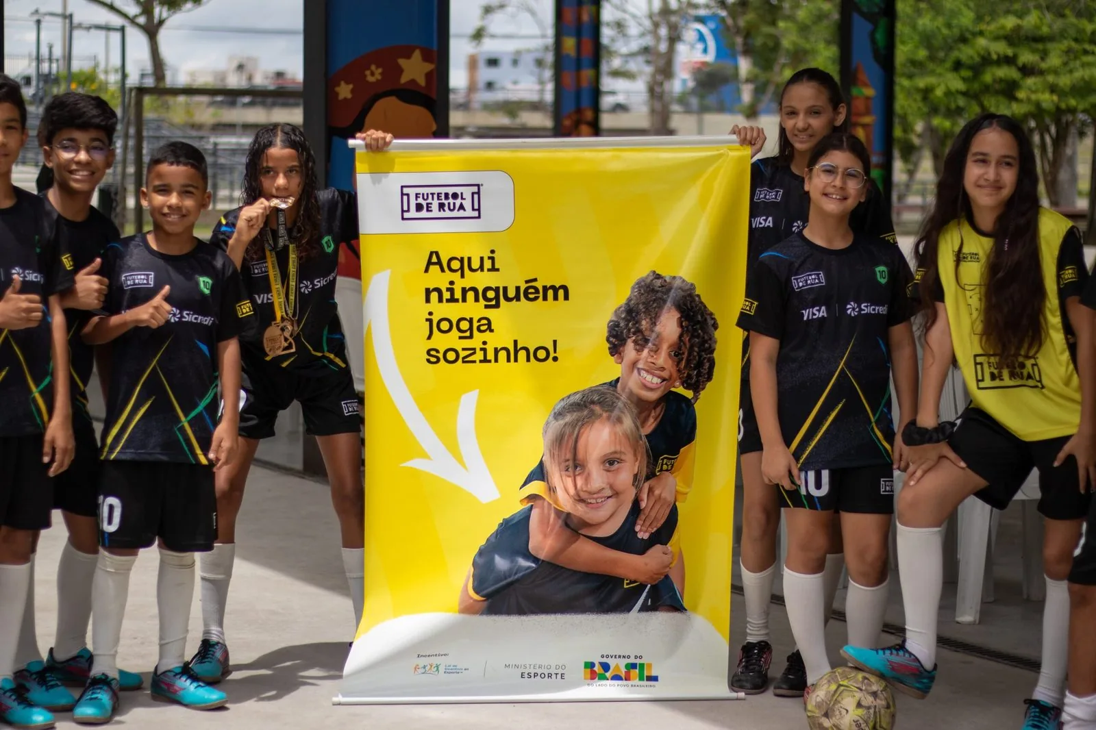 Estudantes com uniformes esportivos ao lado de um banner do projeto Futebol de Rua