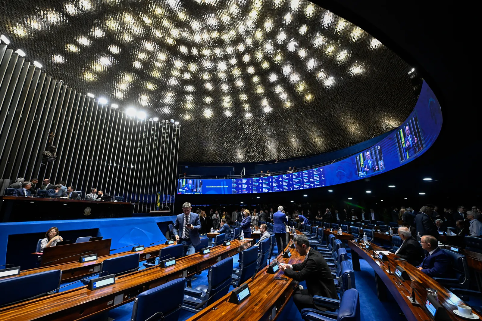 Vista ampla do plenário do Senado Federal, com a mesa diretora, o brasão da República e senadores nas bancadas azuis durante uma sessão.