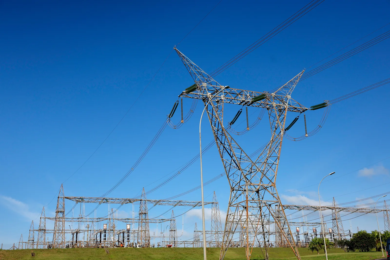 Torre de transmissão de energia elétrica com cabos e isoladores recortada contra céu azul, com um pátio de subestação ao fundo.