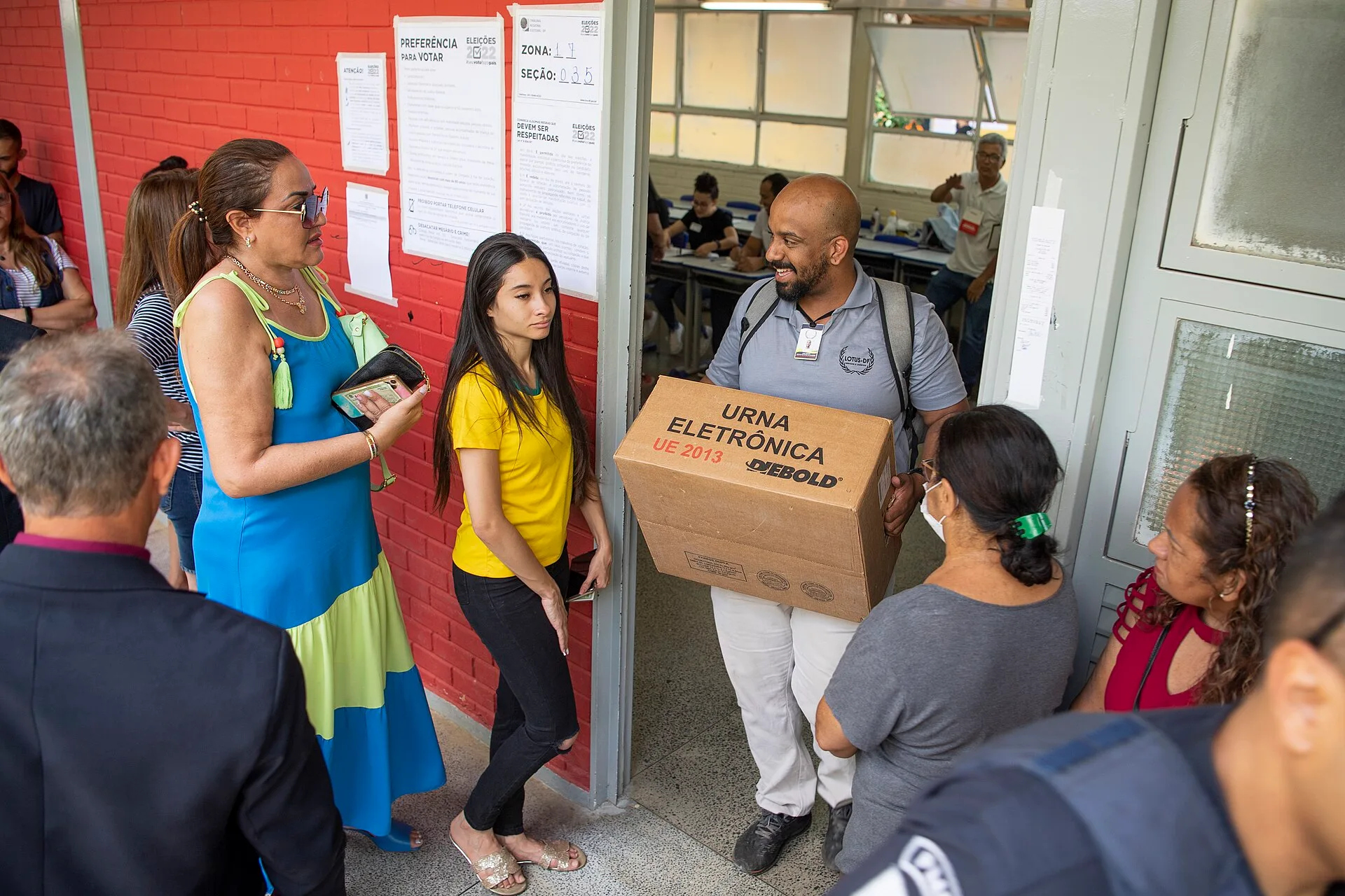 Eleitores conversam na entrada de um local de votação, ao lado de cartazes com as regras da eleição afixados na parede, enquanto um funcionário carrega uma caixa de urna eletrônica.