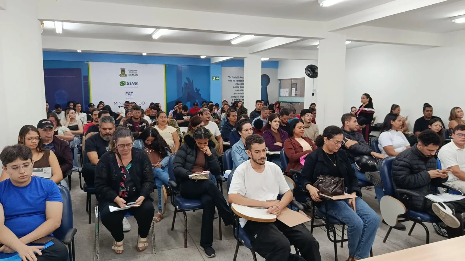 Sala cheia de candidatos sentados em cadeiras escolares, com pranchetas e papéis, diante de um banner do Sine Municipal