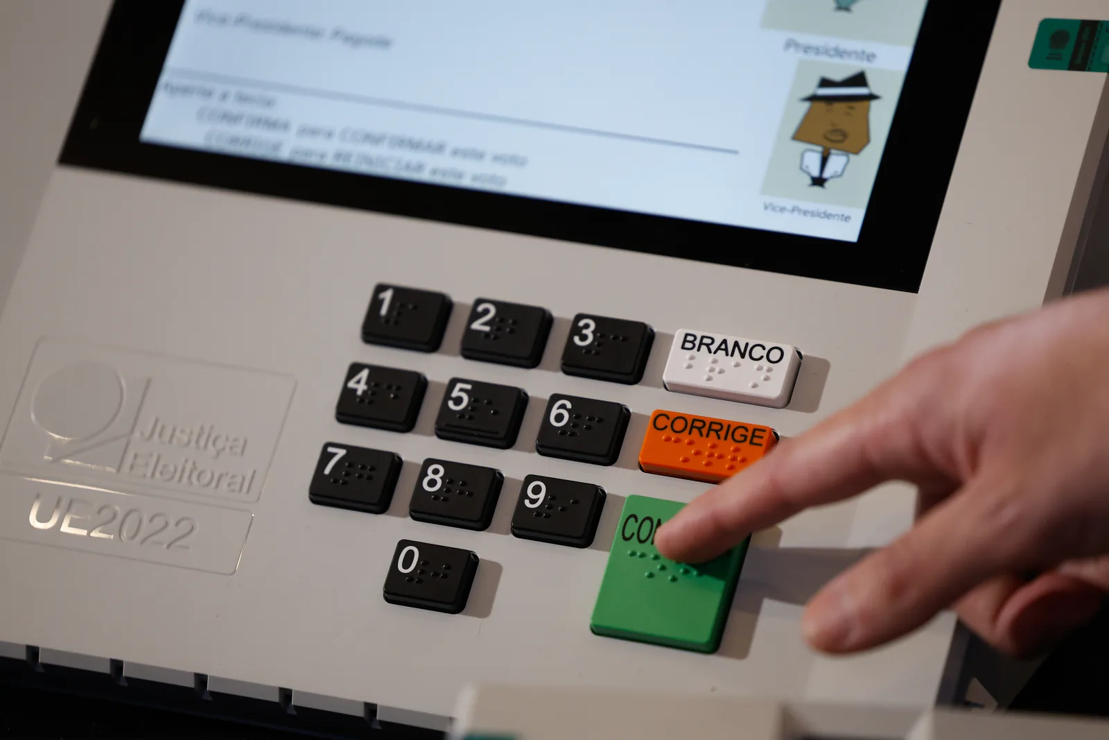 Dedo pressiona a tecla verde de confirmação no teclado numérico de uma urna eletrônica, com a tela exibindo a votação para presidente e vice.