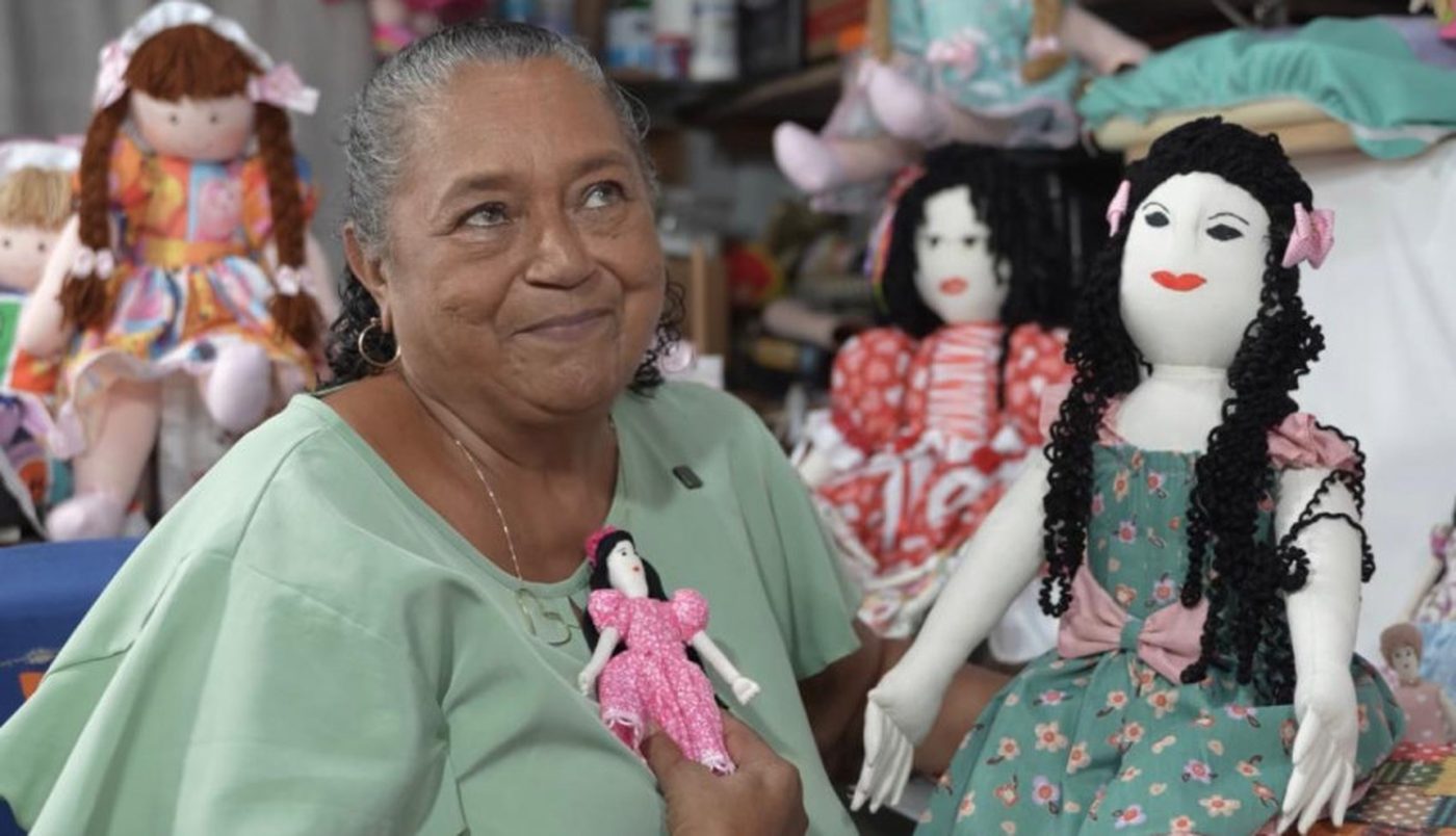 Artesã segura uma boneca de pano ao lado de suas peças no 42º Salão do Artesanato Paraibano, em Campina Grande.