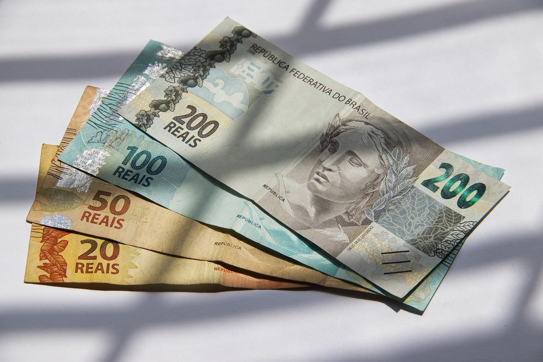 Cédulas de real brasileiras de R$ 20, 50, 100 e 200 dispostas sobre fundo claro.