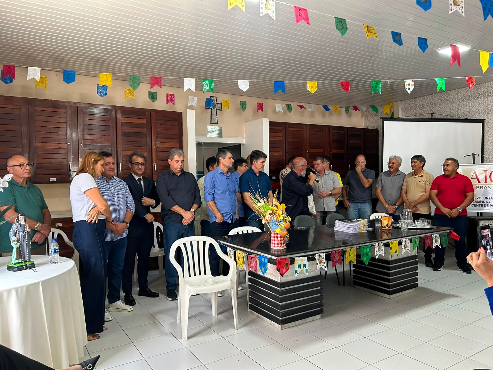 Autoridades e representantes de entidades reunidos em pé em um auditório decorado com bandeirolas juninas, ao lado de uma mesa com pilhas de documentos durante a entrega dos recursos.