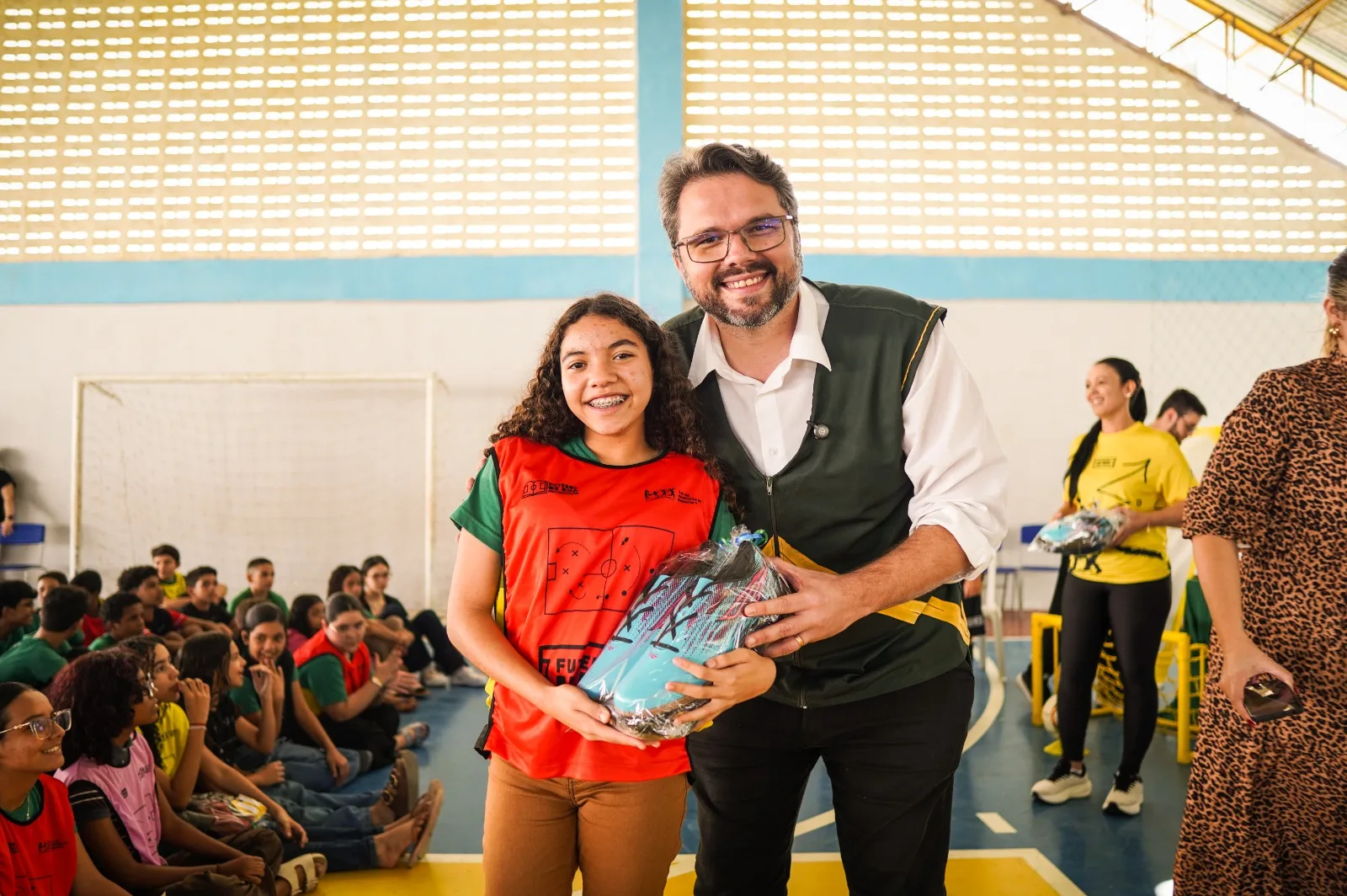 Uma estudante de colete vermelho recebe um kit com chuteiras das mãos de um adulto na quadra da escola, com colegas ao fundo.