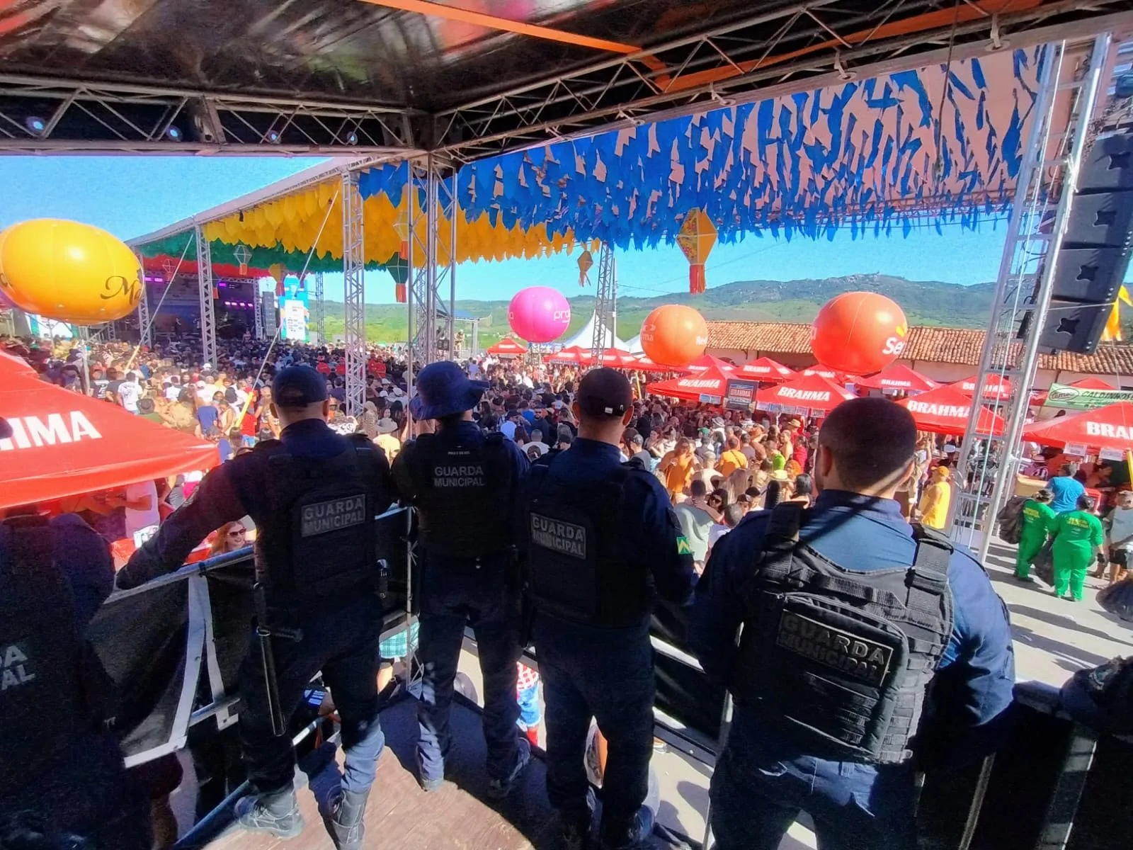 Quatro agentes da Guarda Civil Municipal, de colete e vistos de costas, observam de um ponto elevado a multidão reunida em um polo do São João, com decoração colorida e montanhas ao fundo.