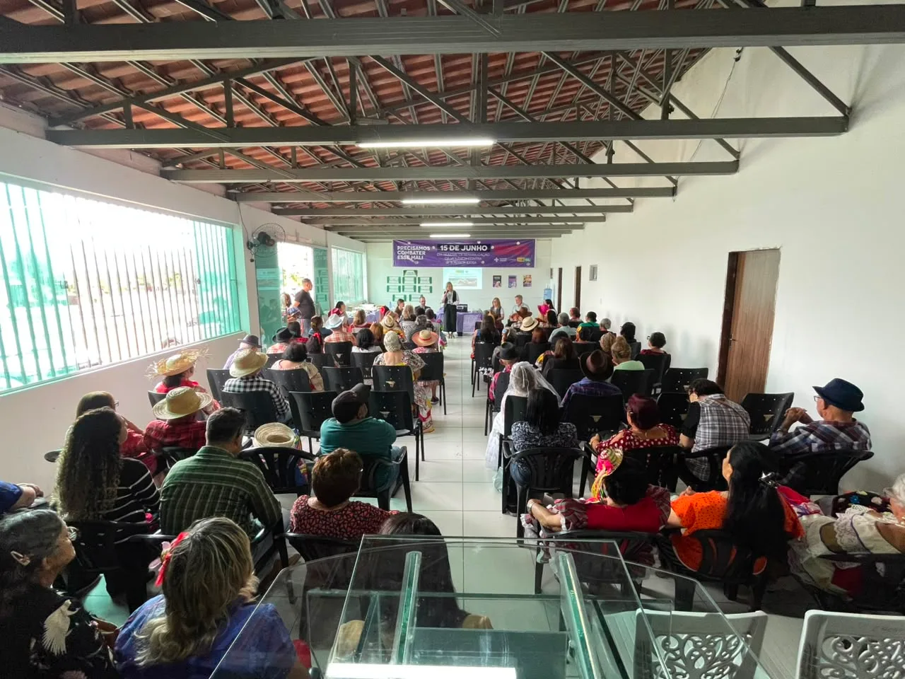 Salão com pessoas idosas sentadas em cadeiras assistindo a uma palestra, sob banner roxo do Dia Mundial contra a violência à pessoa idosa.